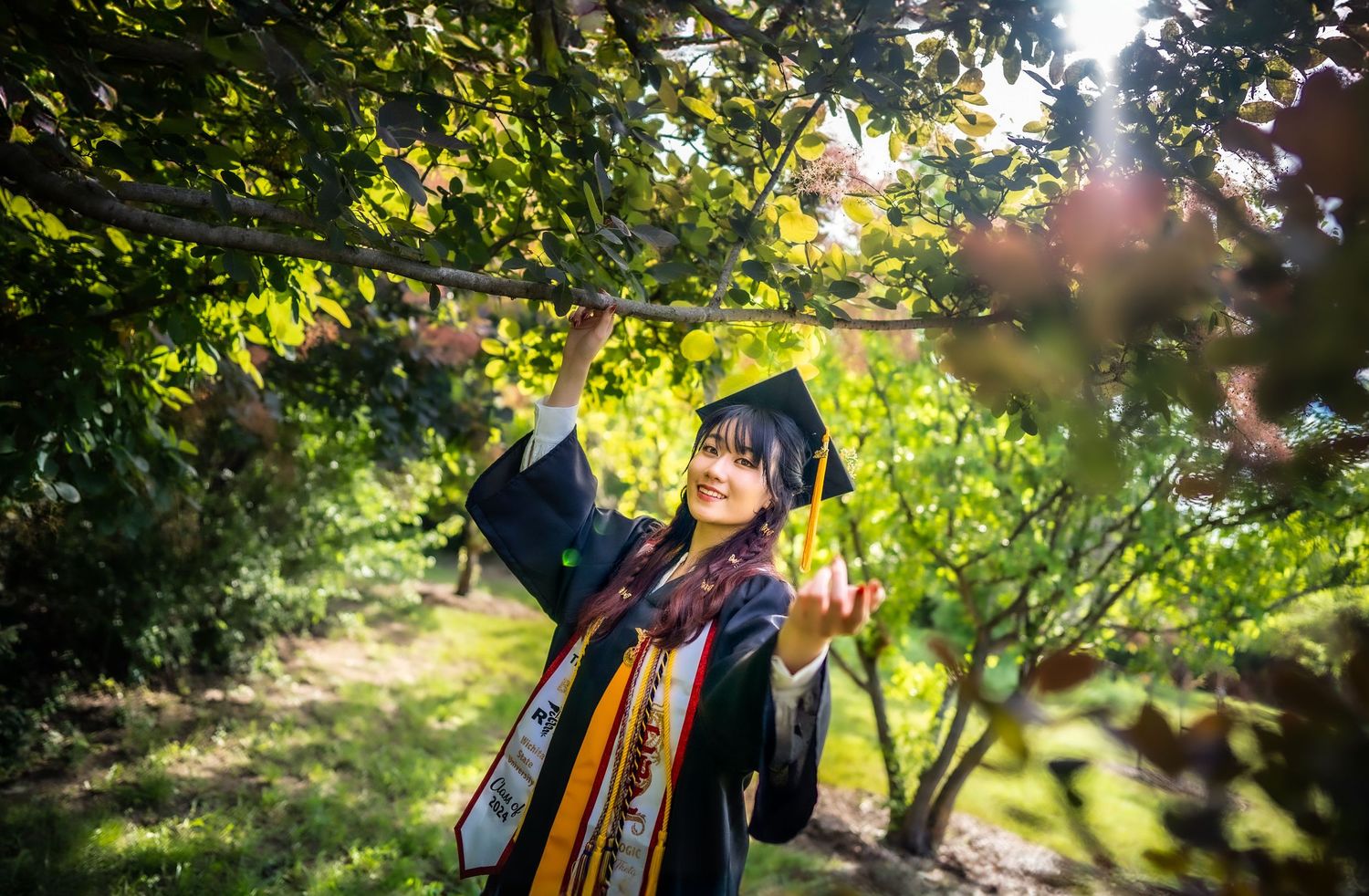 A person in navy blue graduation regalia celebrates outdoors with autumn foliage in the background.