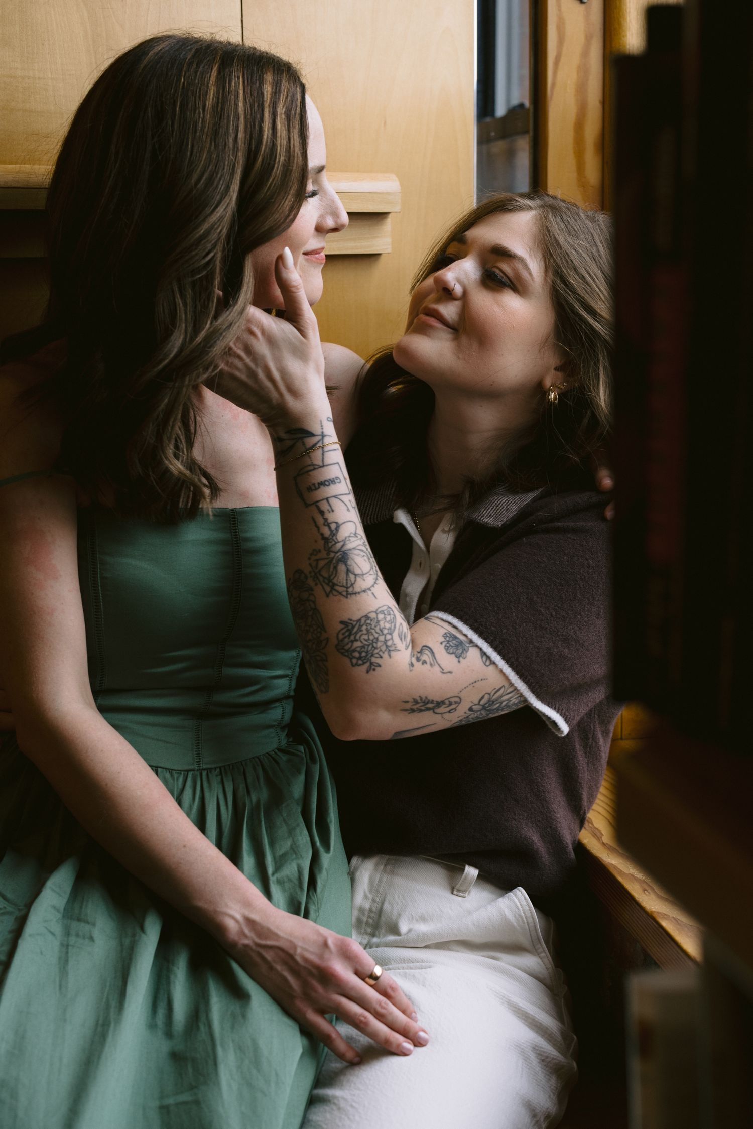 Two women sharing a tender moment, one in a green dress, the other with tattoos in a dark top and white pants.