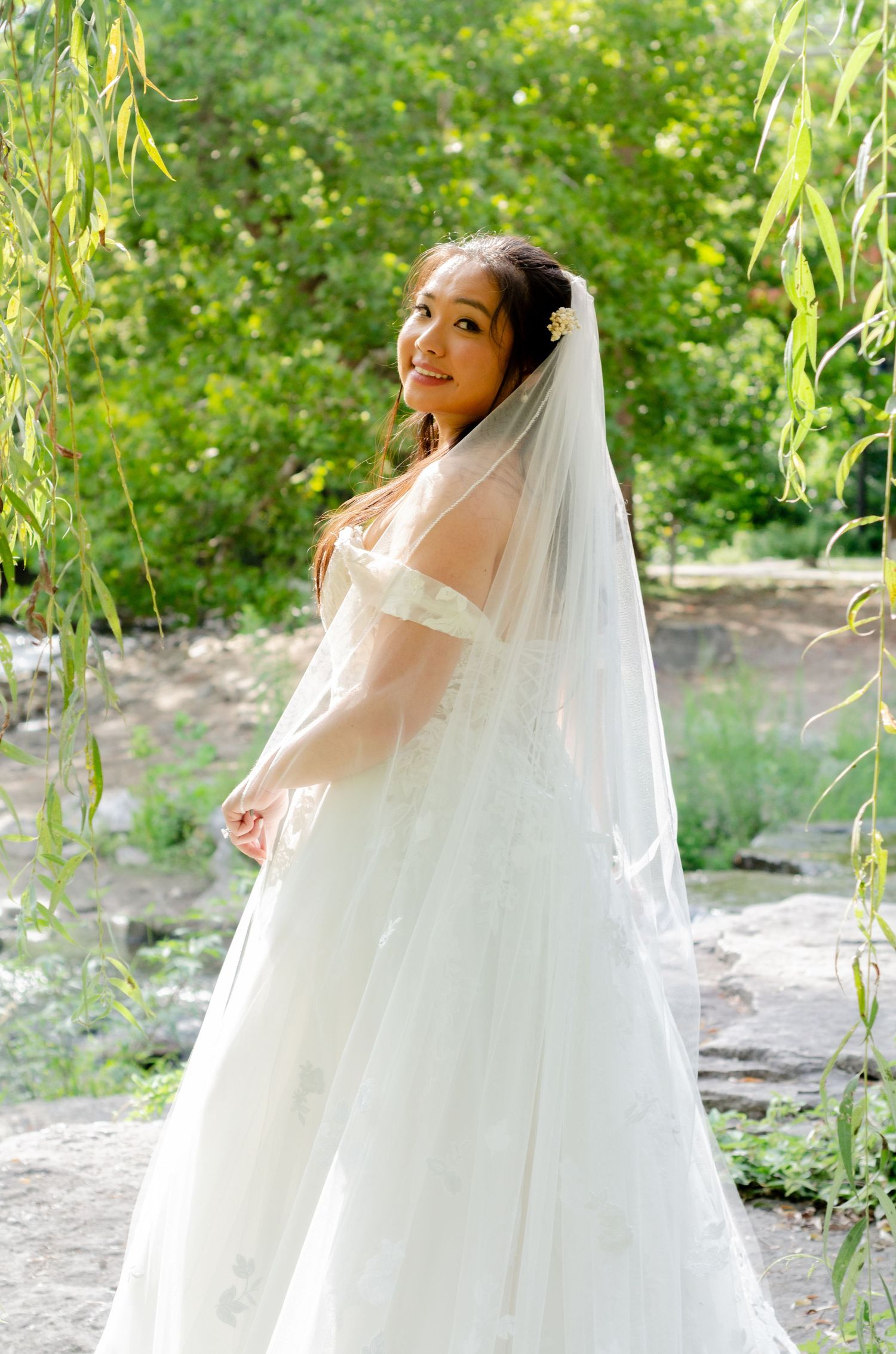 A bride in a flowing white wedding dress poses gracefully in a natural outdoor setting with green foliage.