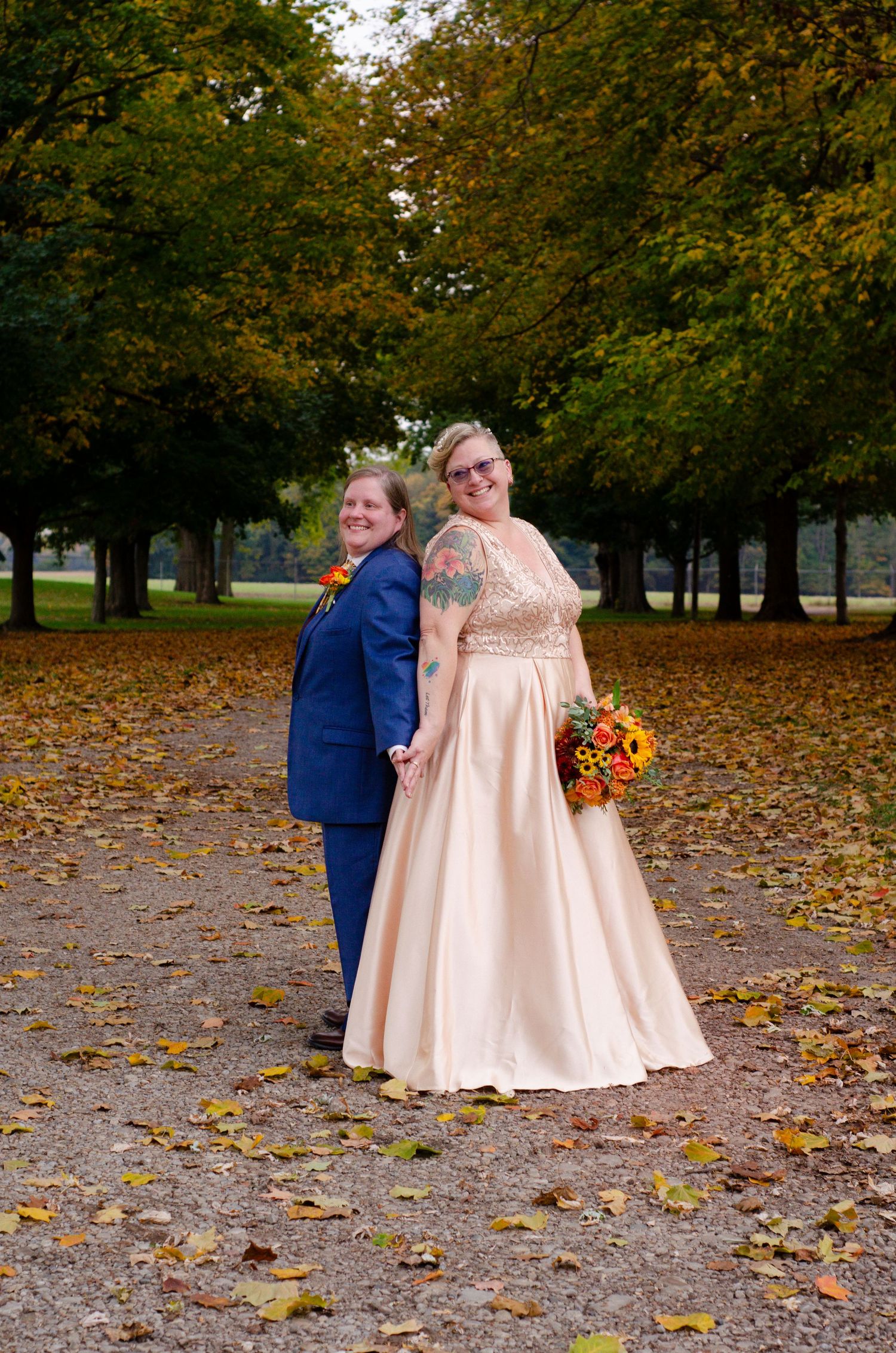 A couple in wedding attire walks through autumn leaves in a park setting with trees in the background.