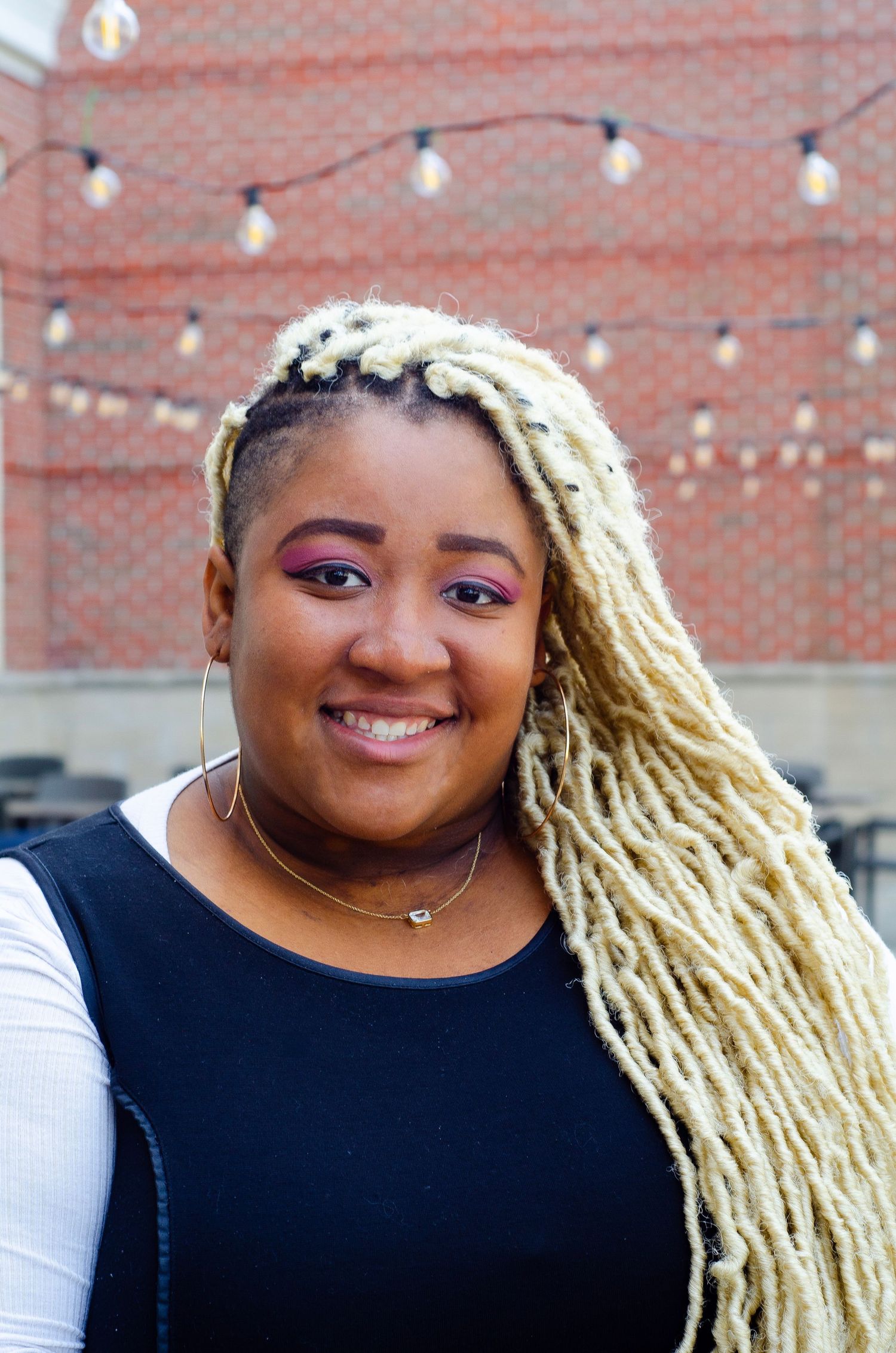 Portrait featuring blonde dreadlocks and black top against brick wall with string lights.
