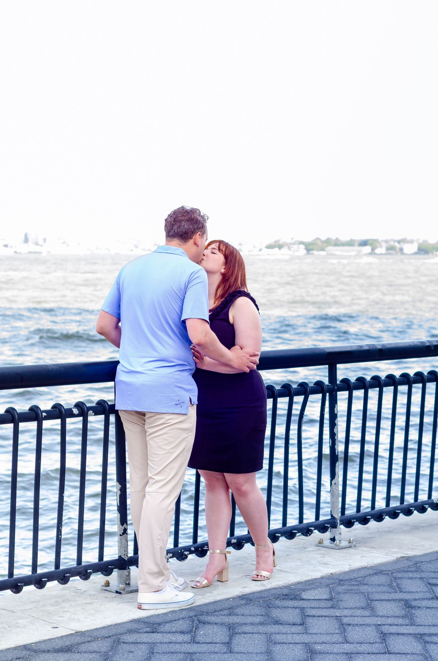 Couple sharing a romantic moment along a waterfront railing with city skyline in background. Engagement photos by KM Conceptions Photography
