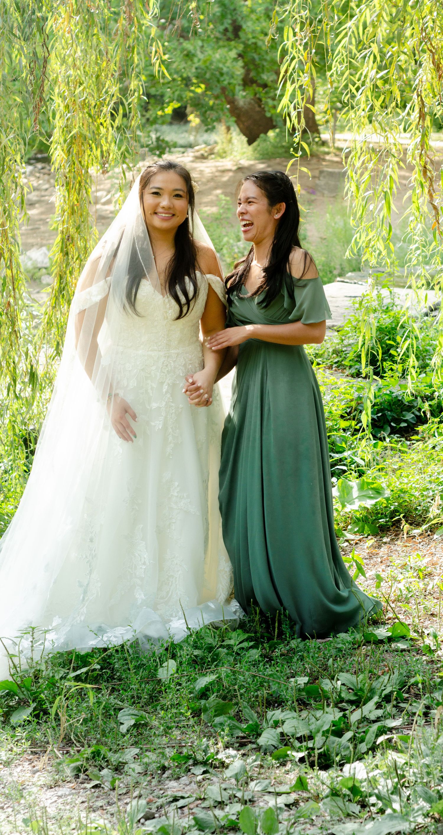 Two people in white and green dresses share a joyful moment under a weeping willow tree in a garden setting.