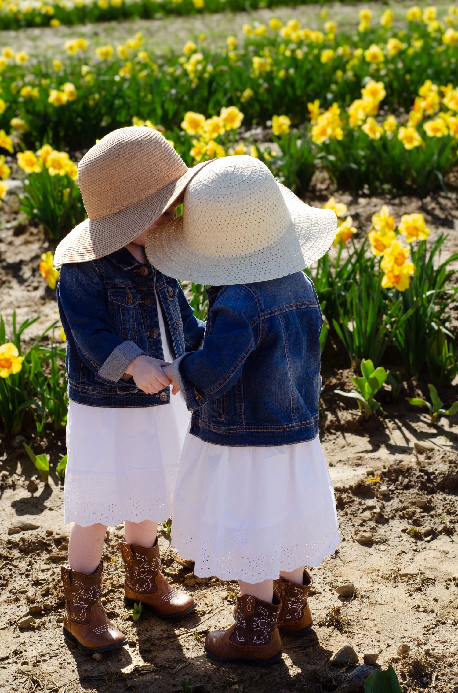 Two small figures in matching denim jackets, white dresses and sun hats stand among blooming yellow daffodils in a garden.