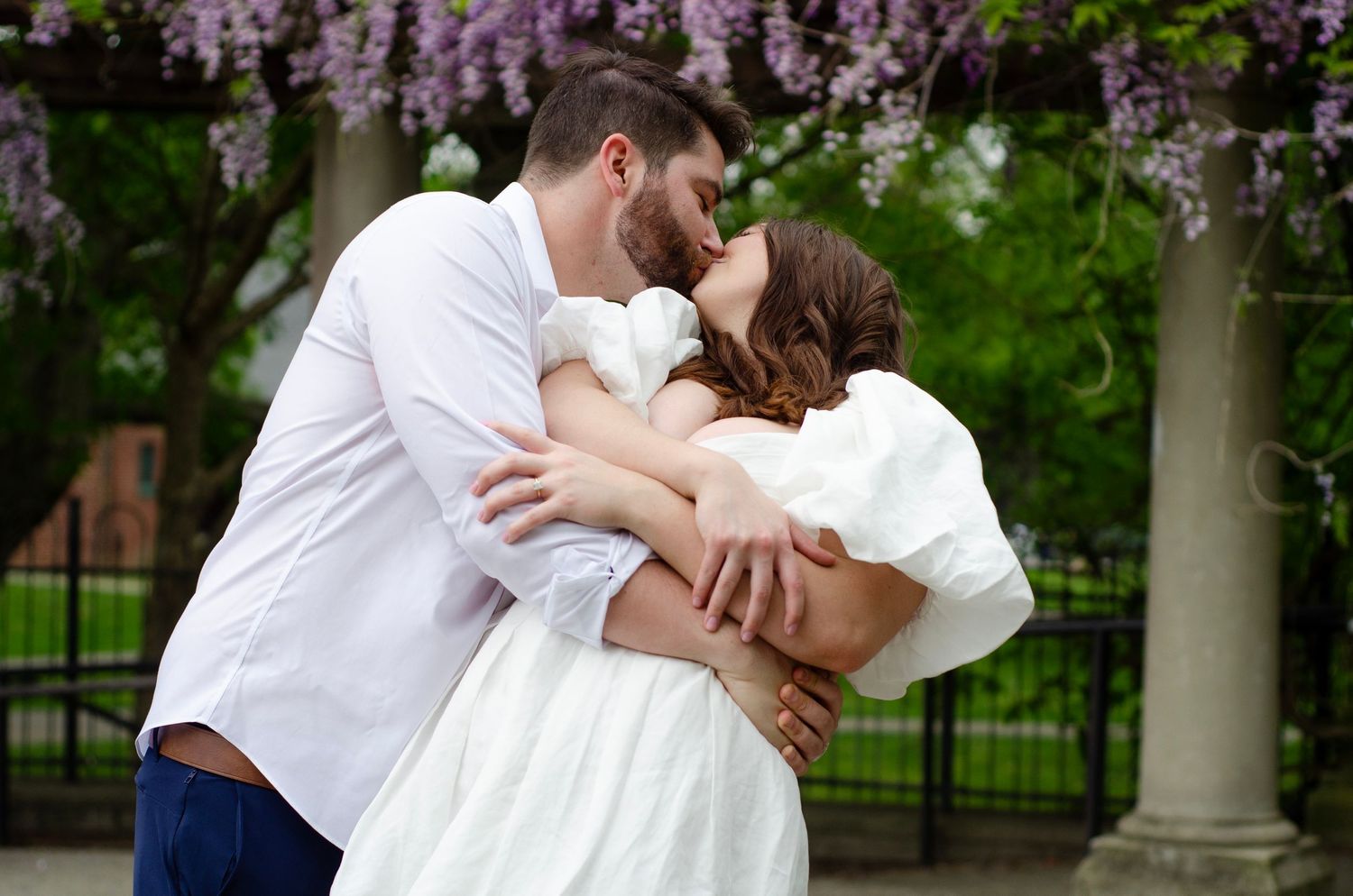 Romantic couple sharing an intimate dip kiss under blooming purple wisteria in a garden setting.