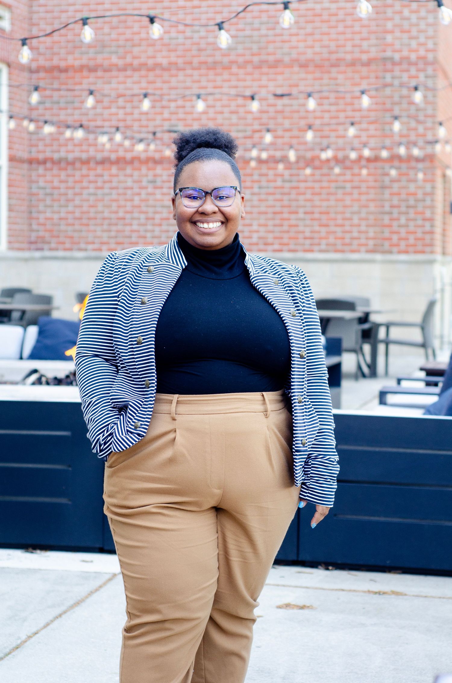 Portrait showing striped blazer and khaki pants against brick wall with string lights.