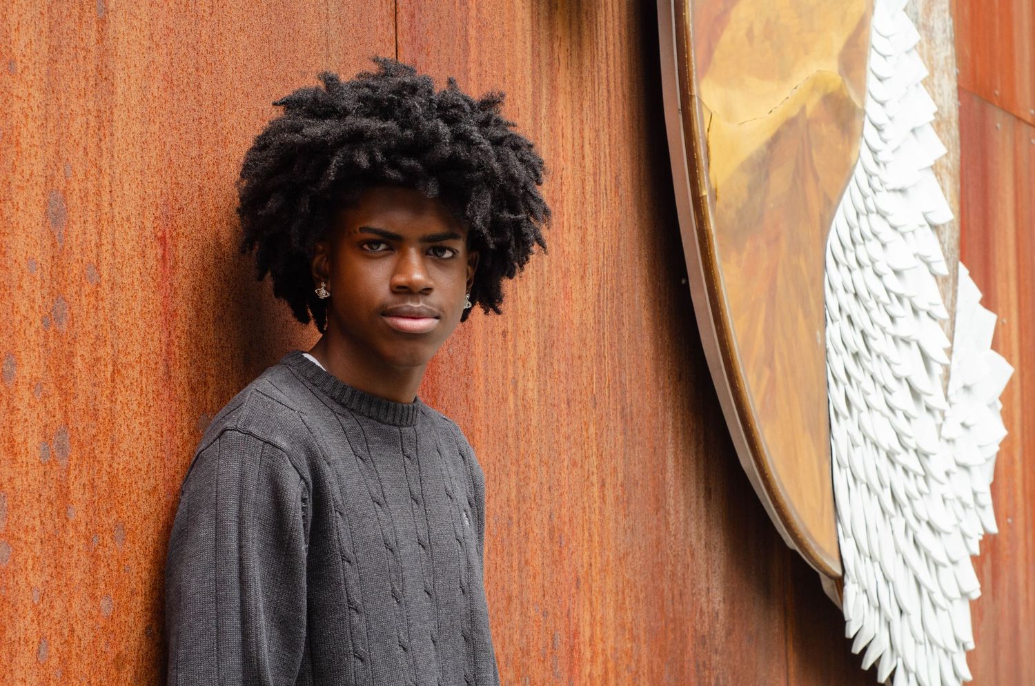 Person in gray sweater poses against rustic orange wall near decorative wing artwork.