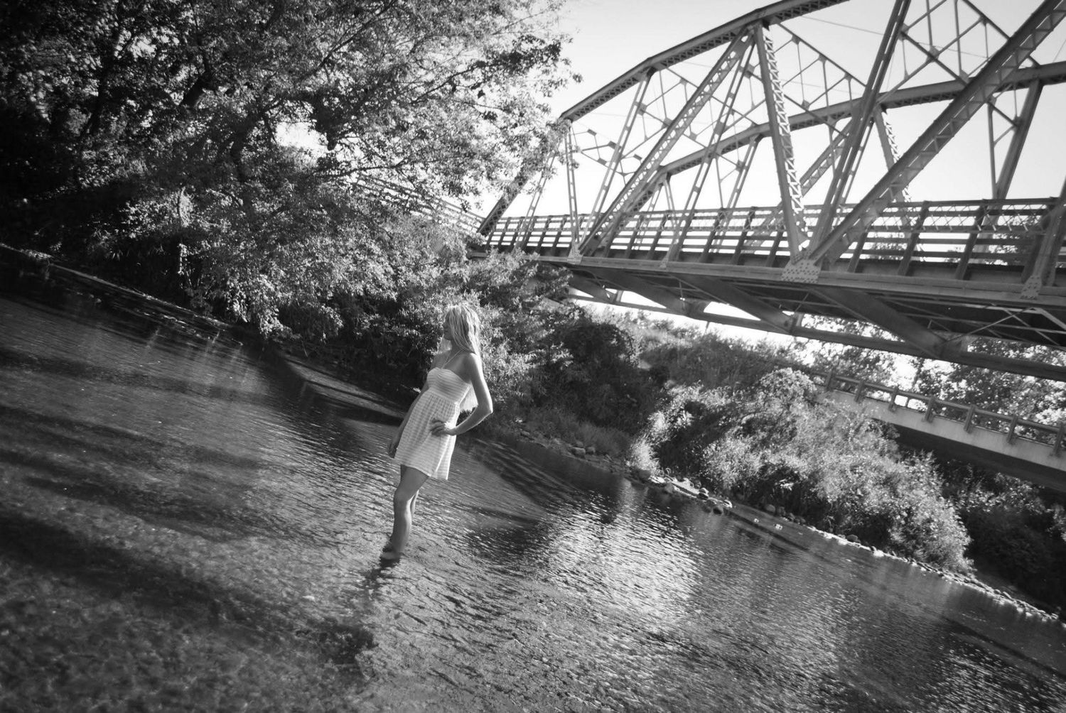 A rustic steel bridge spans over a peaceful river surrounded by lush green trees and vegetation.