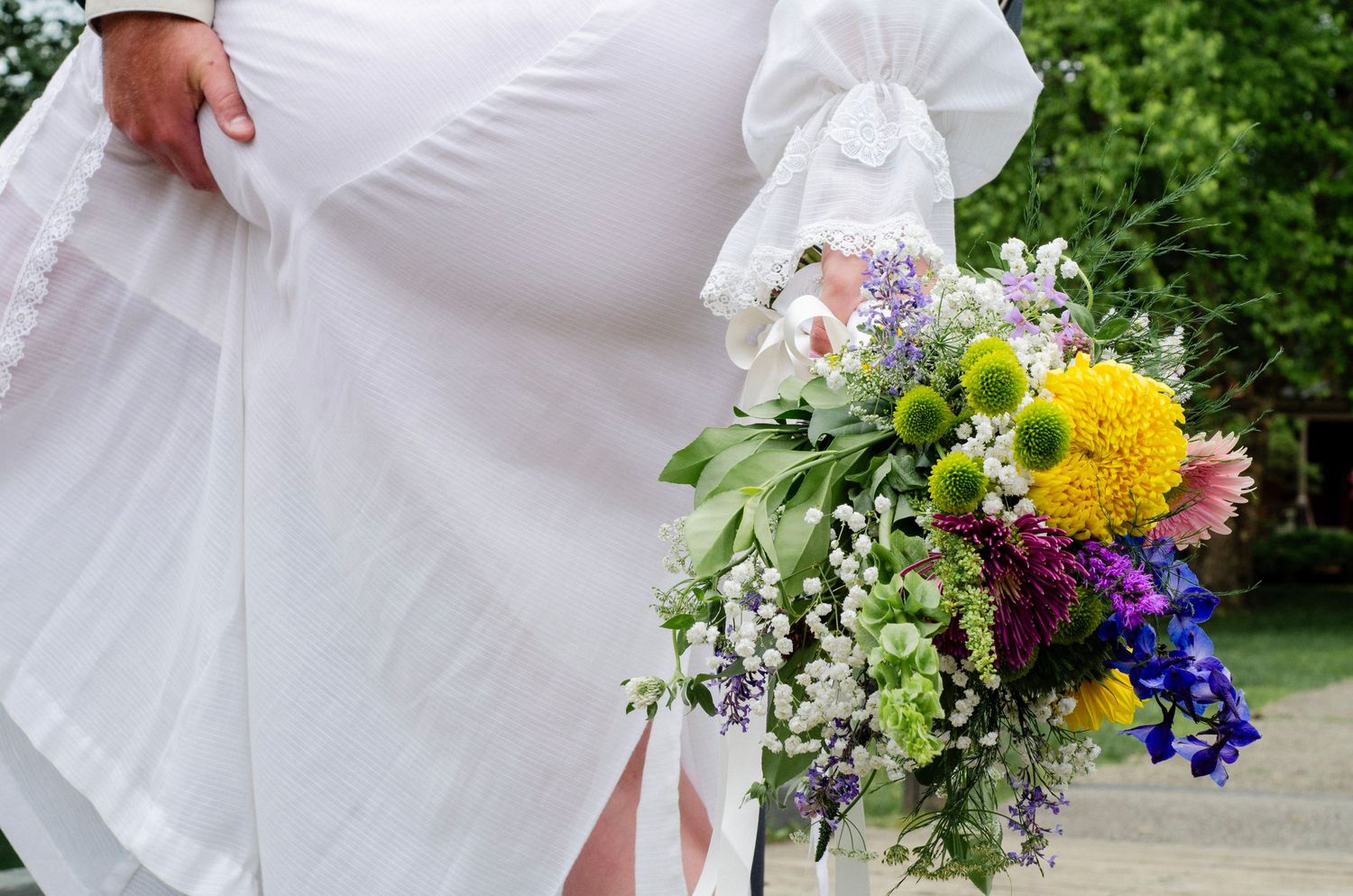 A colorful wedding bouquet with yellow flowers, purple flowers, and greenery against a white dress. Wedding photos by KM Conceptions.