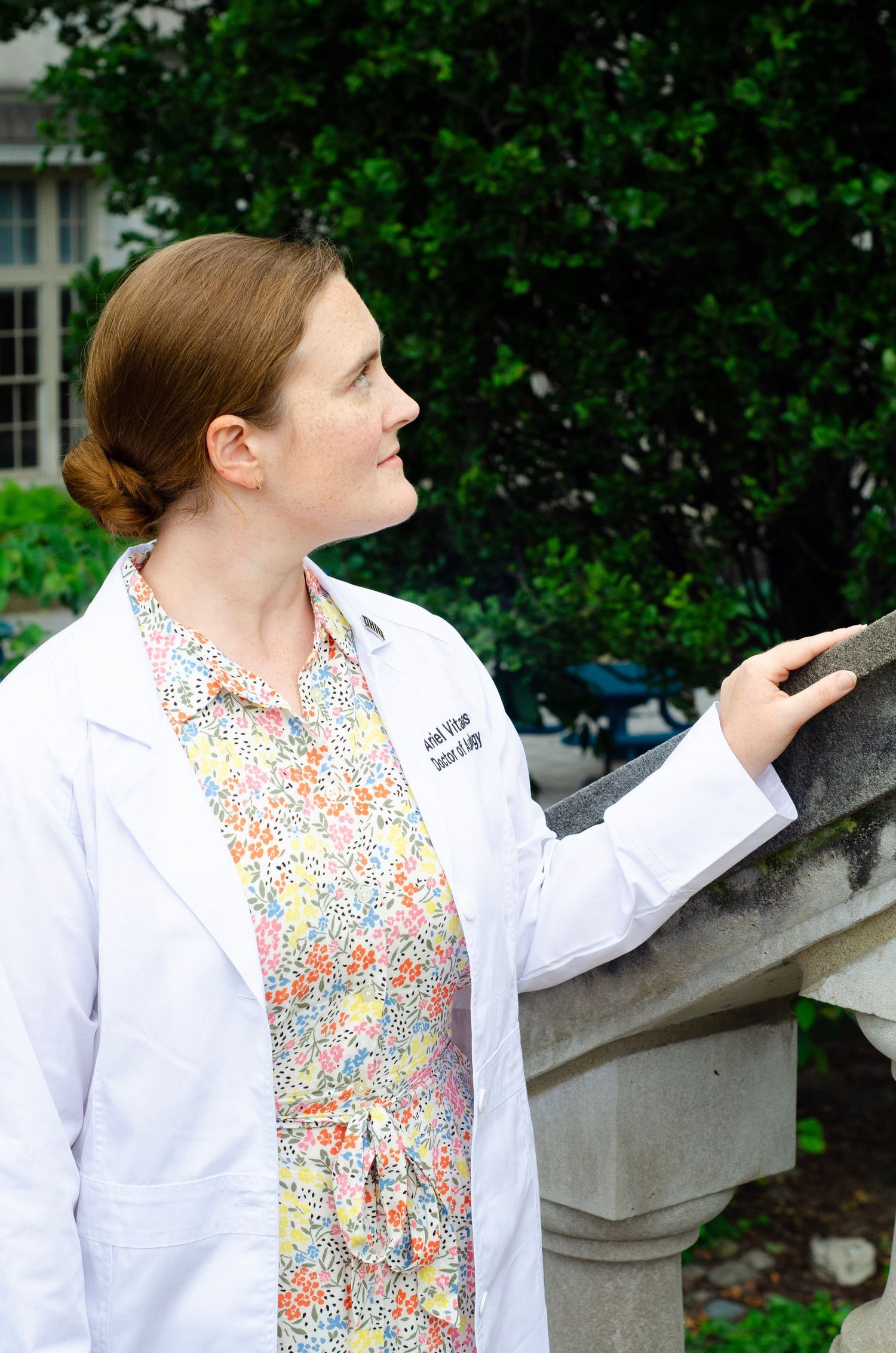 Student in white lab coat and floral dress stands by stone railing on campus.