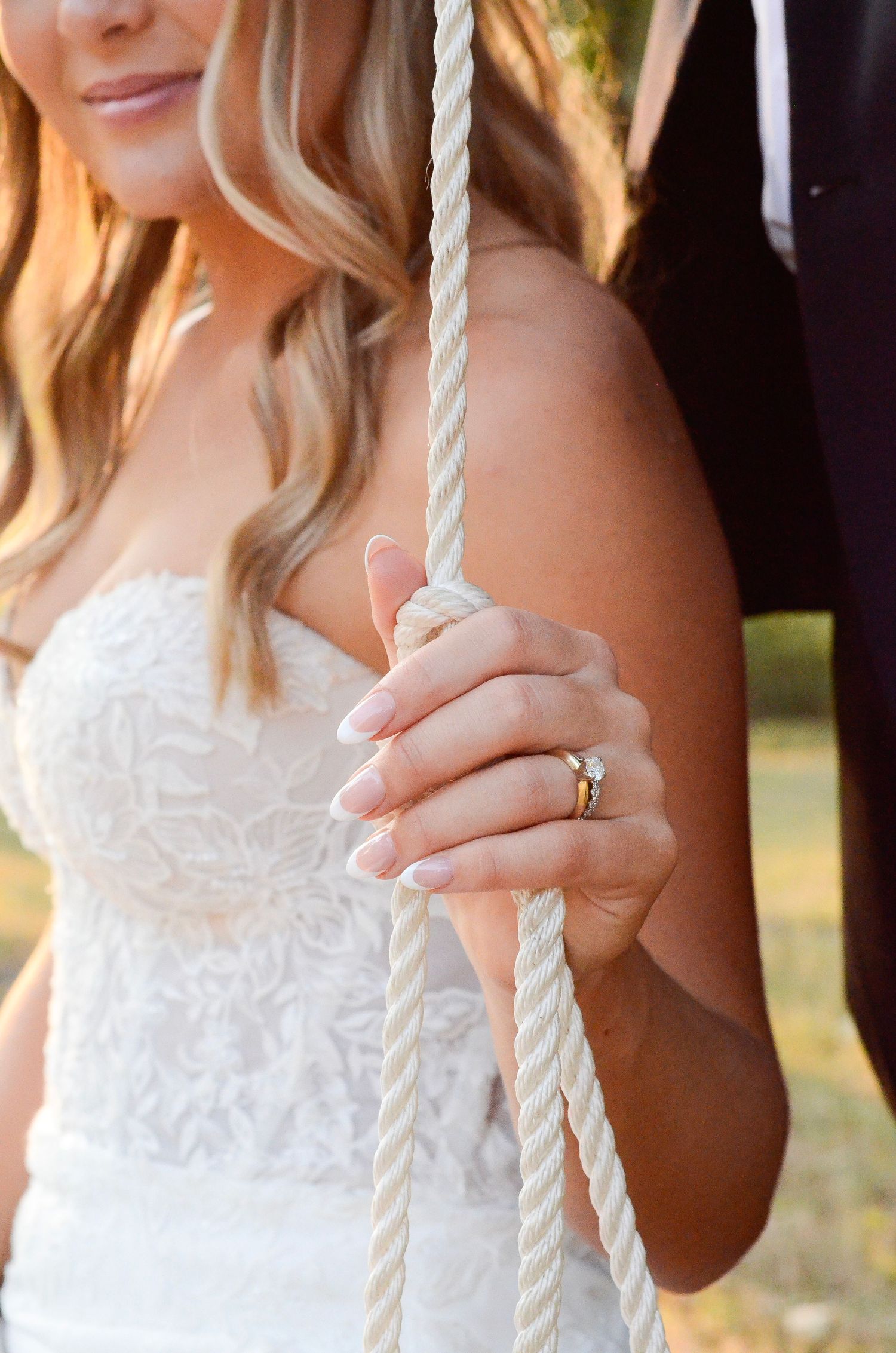 Close-up of a bride in a white lace dress holding a rope swing, showing her wedding ring in golden sunlight. Wedding photos by KM Conception