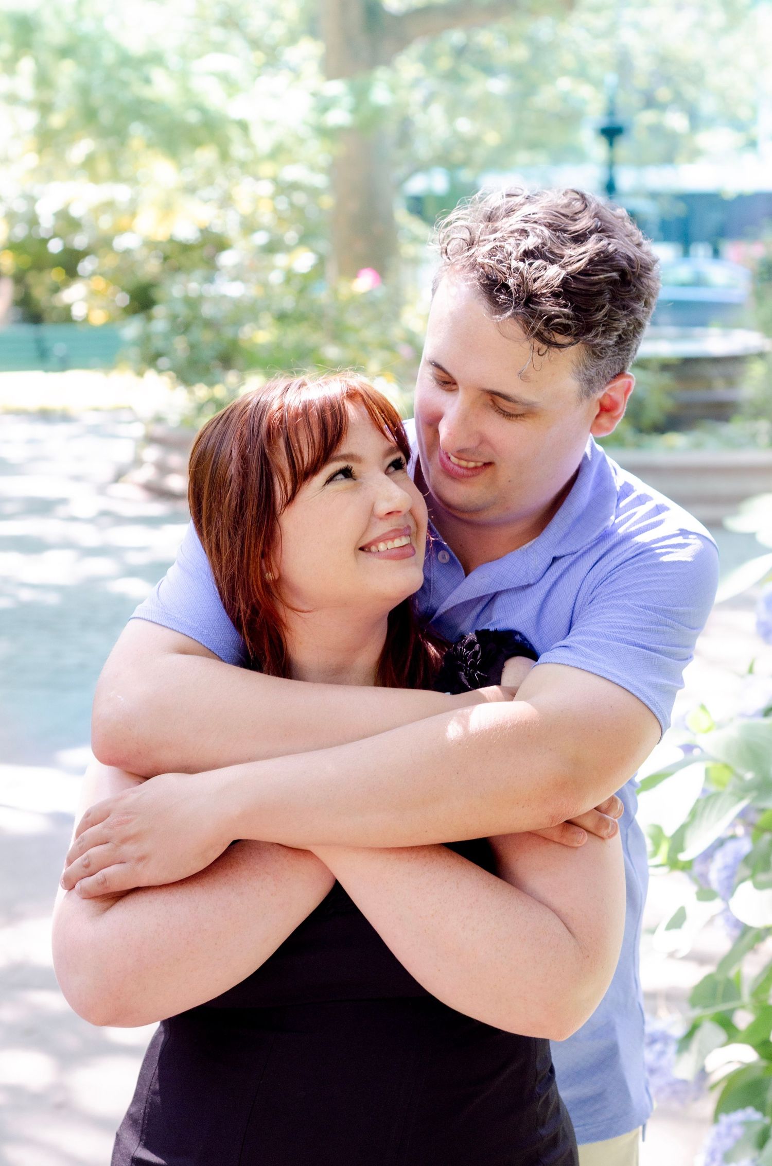 A smiling couple embraces outdoors, woman with red hair looks up lovingly at curly-haired man in blue polo shirt.