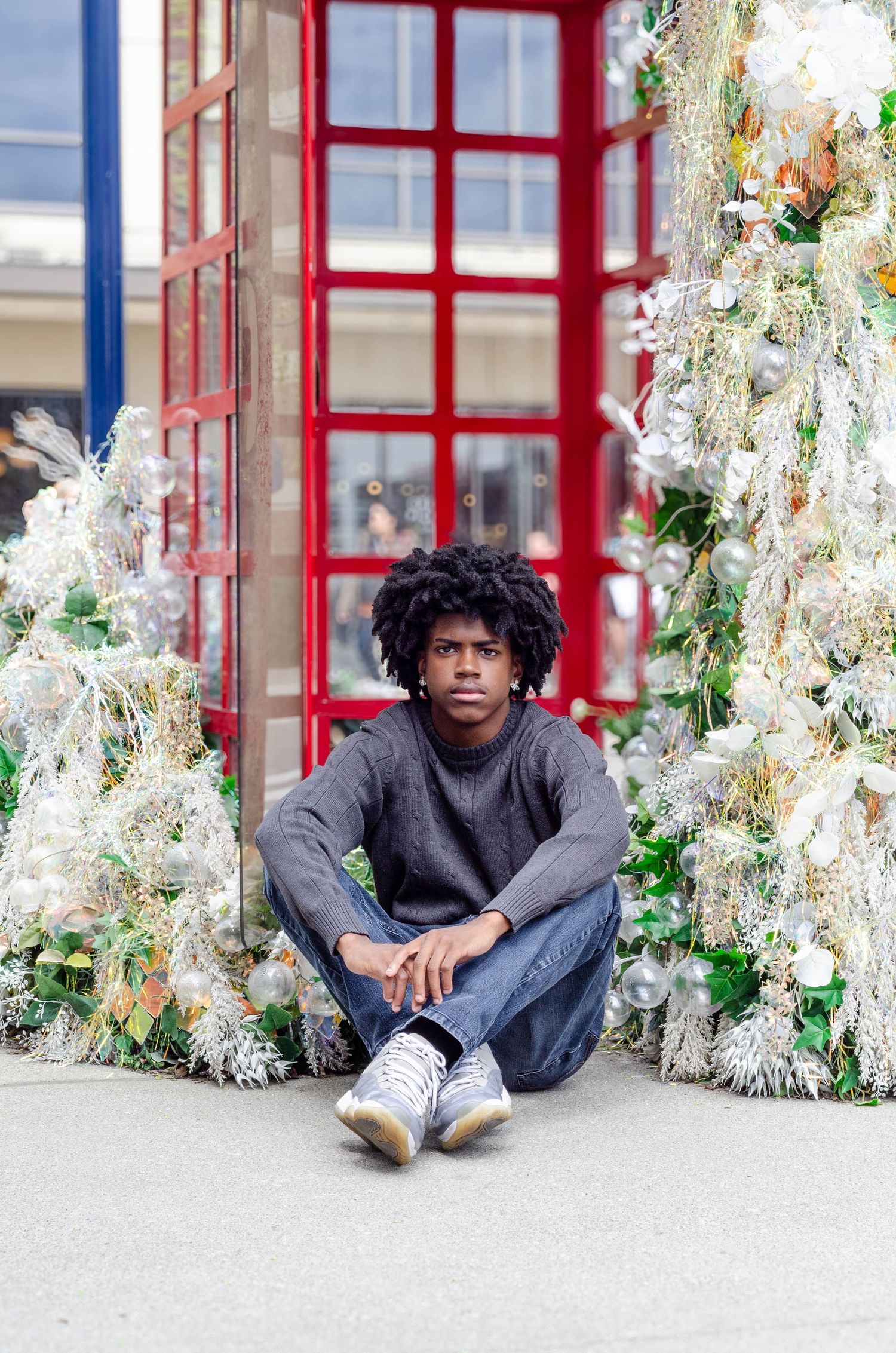 Person sits cross-legged surrounded by white tinsel and holiday decorations near red window.