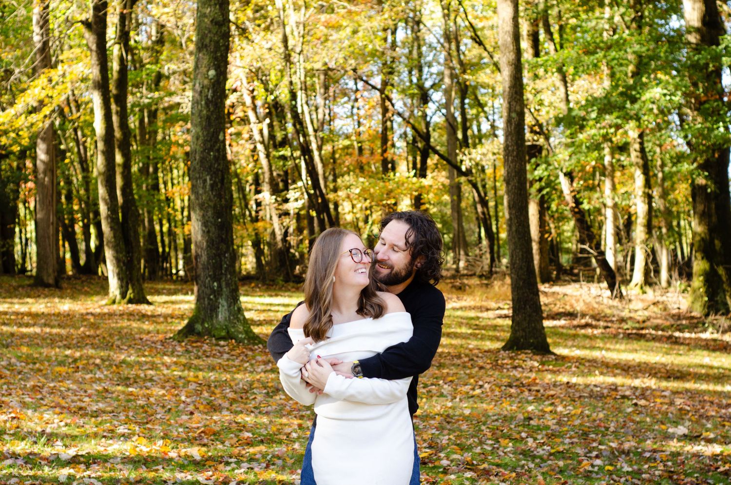 Couple embracing and smiling in autumn forest with colorful fall foliage and leaf-covered ground.