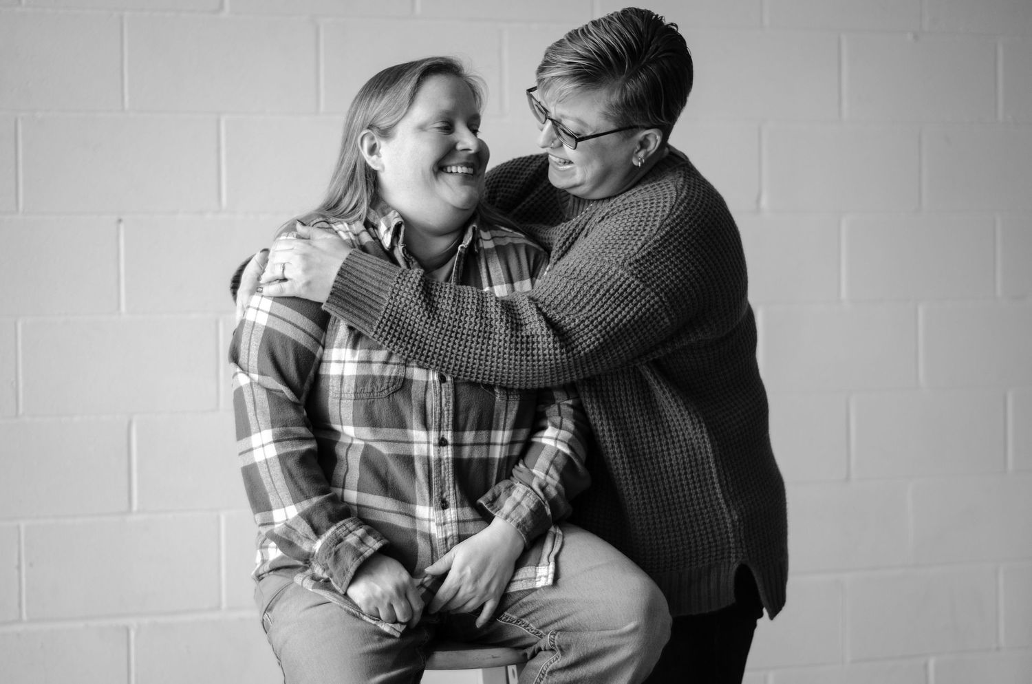 Black and white portrait of a couple embracing in casual attire against a white wall. Engagement photos by KM Conceptions Photography