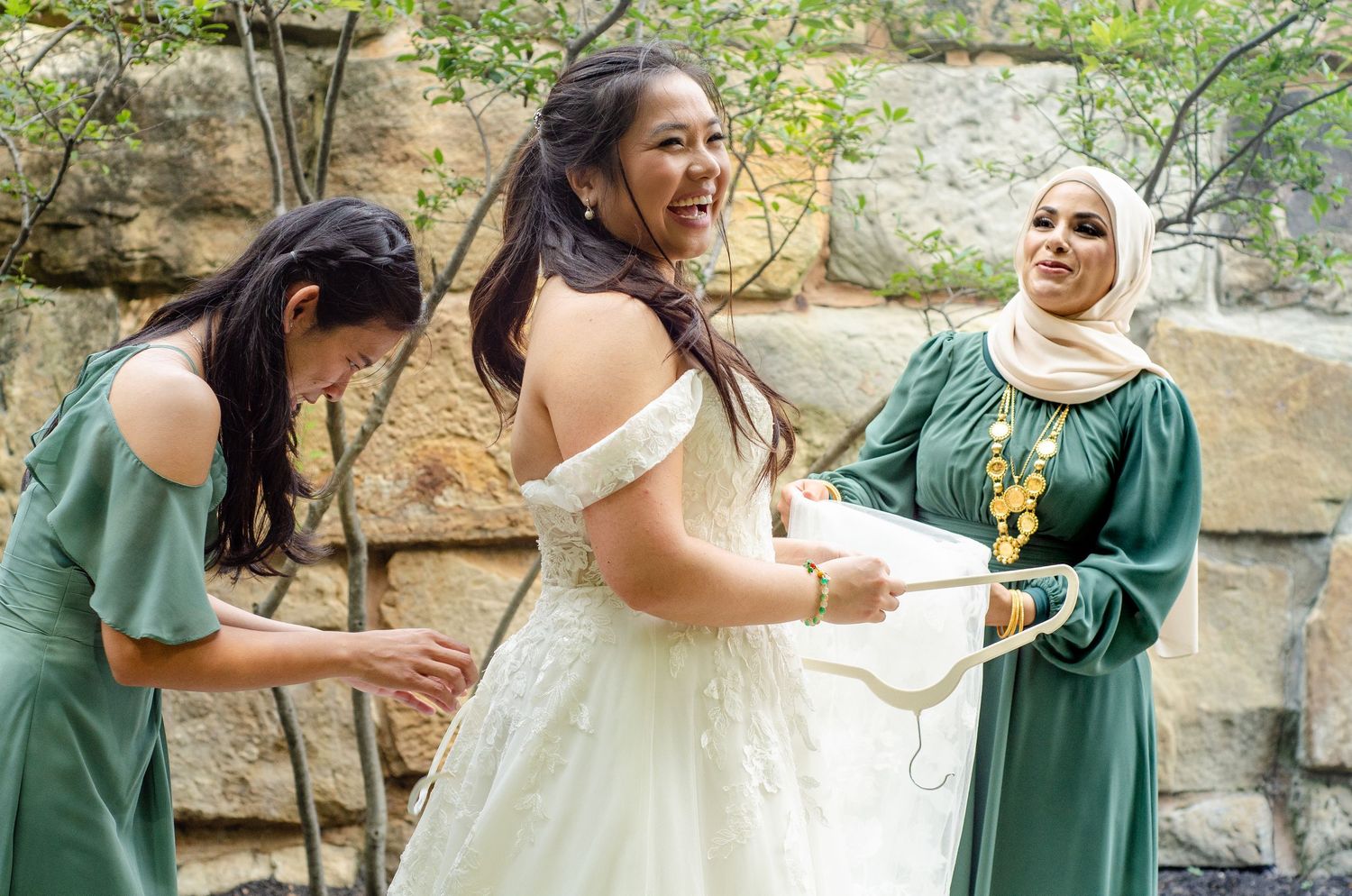 Wedding party in emerald green dresses poses together outdoors against stone wall backdrop.