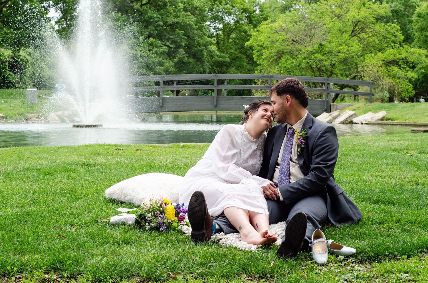 Couple in wedding attire sitting on grass by a fountain and bridge in a lush green park on their wedding day.