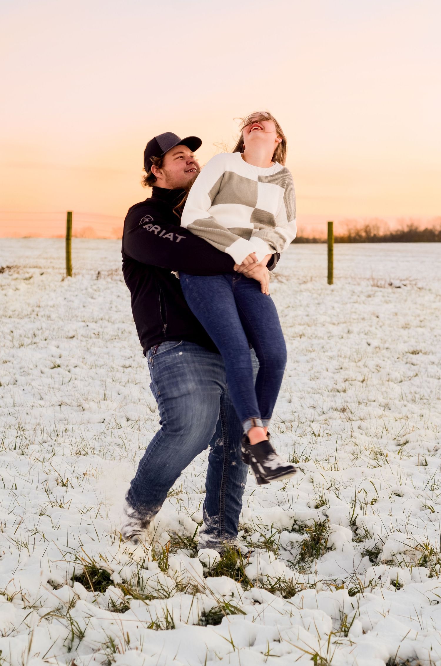A playful couple dances and embraces in a snow-covered field at sunset with fence posts visible in the background.