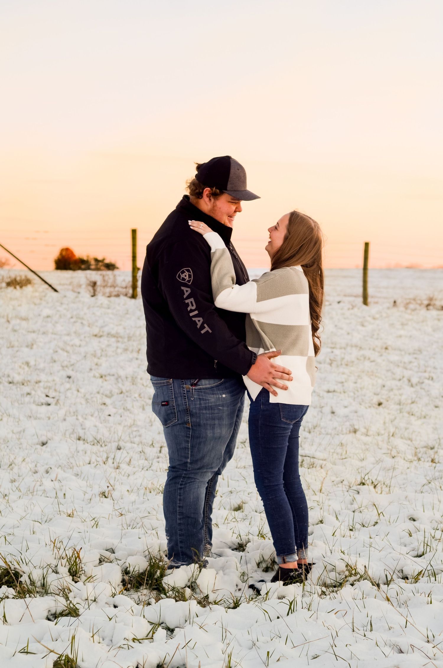 Couple embracing in a snowy field during sunset while wearing casual winter outfits.