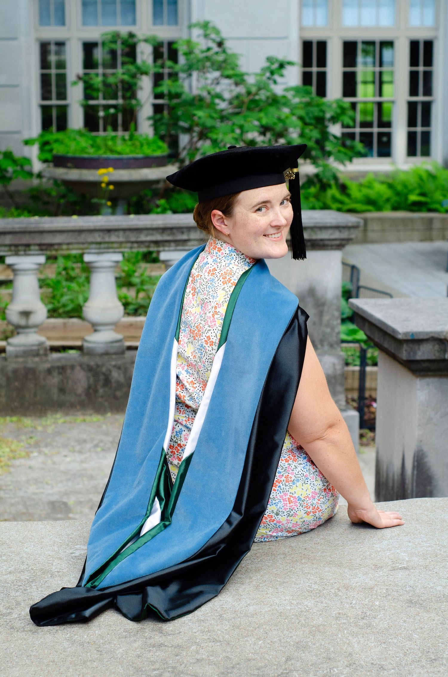 Graduate in light blue academic regalia sits on stone steps during graduation photo session.