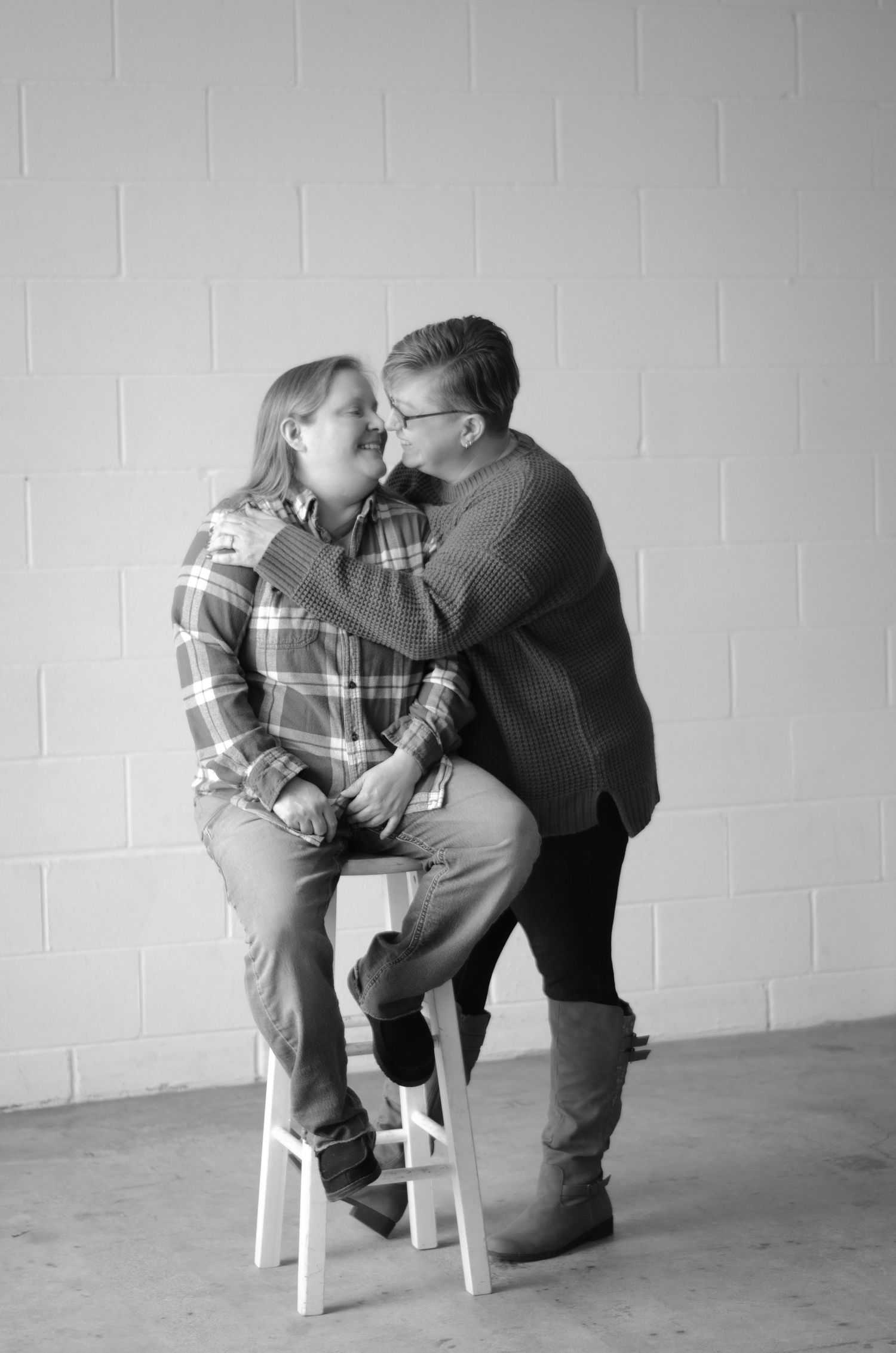 Black and white portrait of couple sharing intimate moment while seated on white stool.
