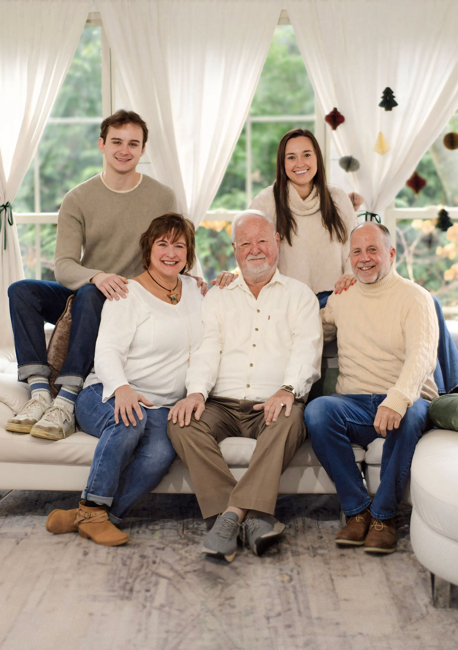 Family members gather on a white couch for a portrait with sheer curtains and holiday decorations behind them.