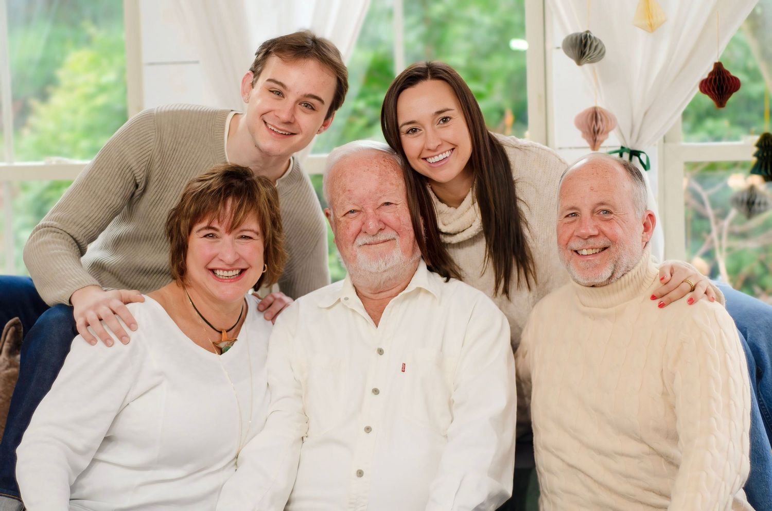 Multi-generational family poses together on a couch with sunlit windows and holiday decorations in background.