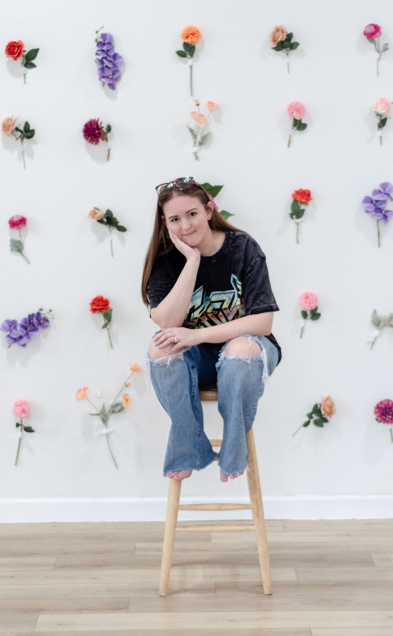 Young woman sitting on wooden stool against white wall decorated with colorful artificial flowers, wearing ripped jeans and graphic tee.