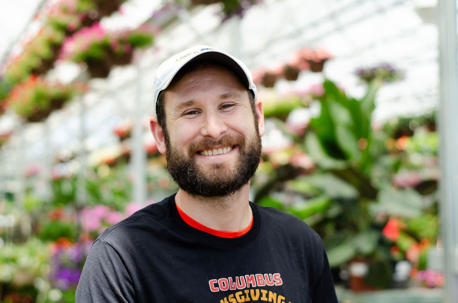 A greenhouse worker in a black t-shirt and baseball cap surrounded by colorful flowers.