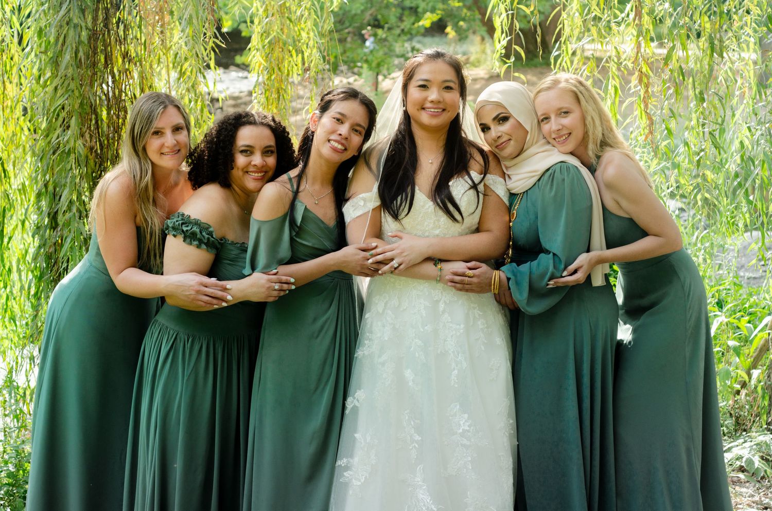 Wedding party in matching emerald green bridesmaid dresses pose with bride in white gown under weeping willow tree.