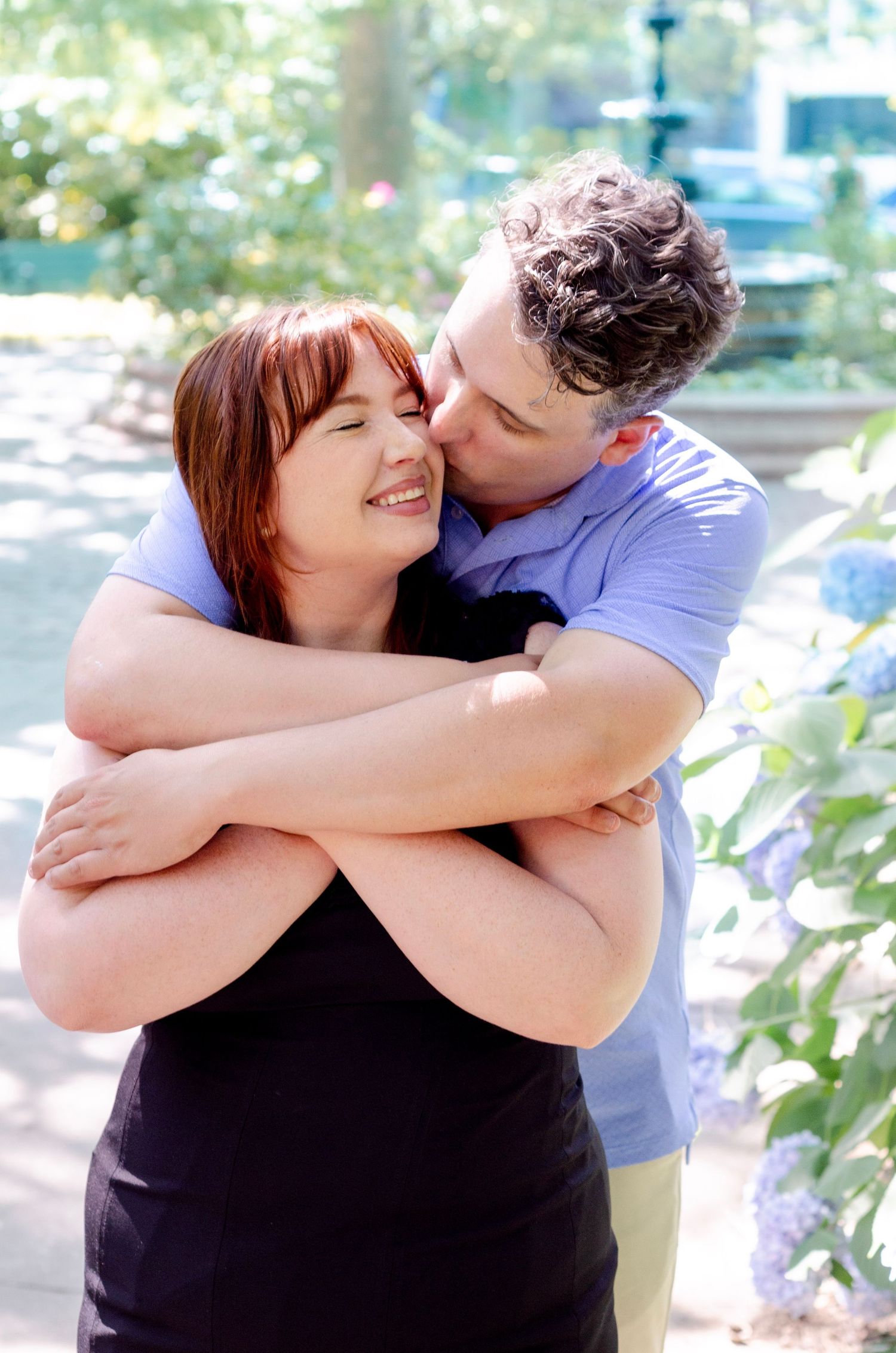 A couple embraces outdoors, man kissing woman's cheek near blooming hydrangeas in a sunny garden setting.
