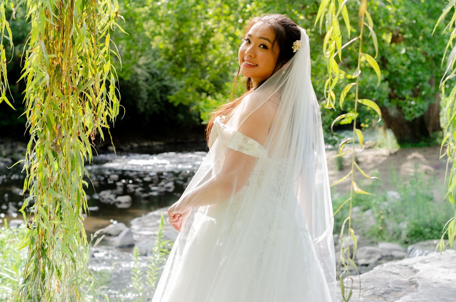 A bride in a white wedding dress stands under hanging willow branches in a garden setting.