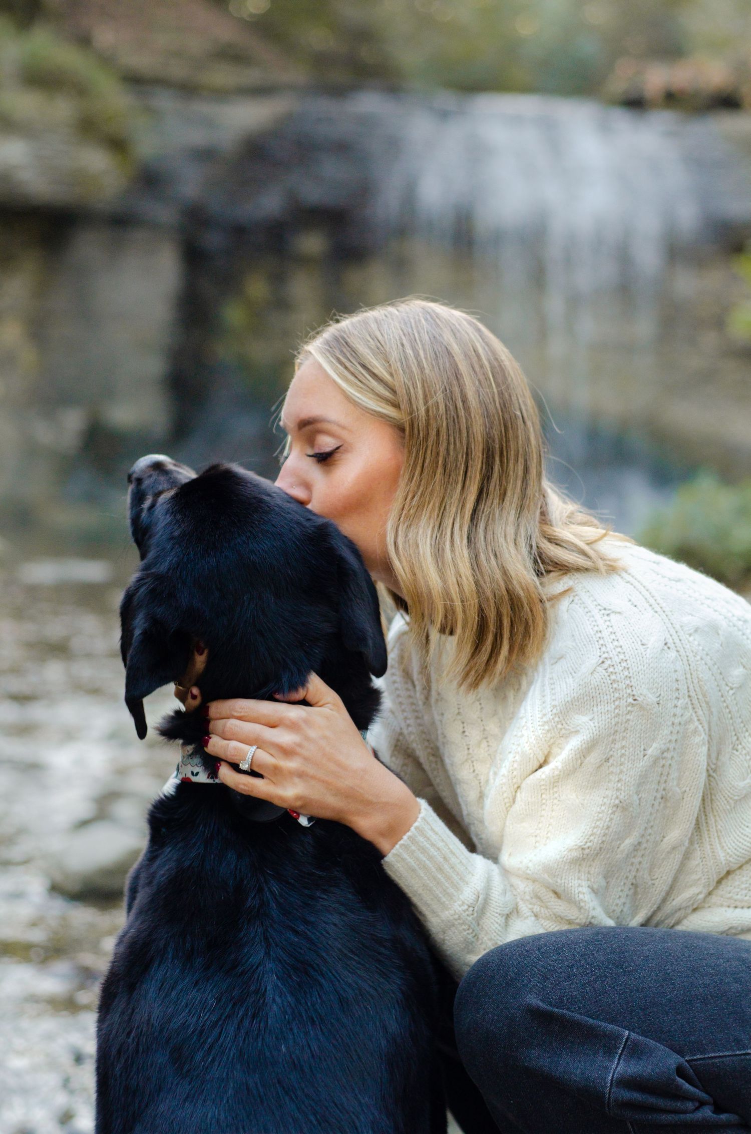 Person in white sweater showing affection to a black dog by a stone wall. Waterfall session photography by KM Conceptions in Columbus, OH.