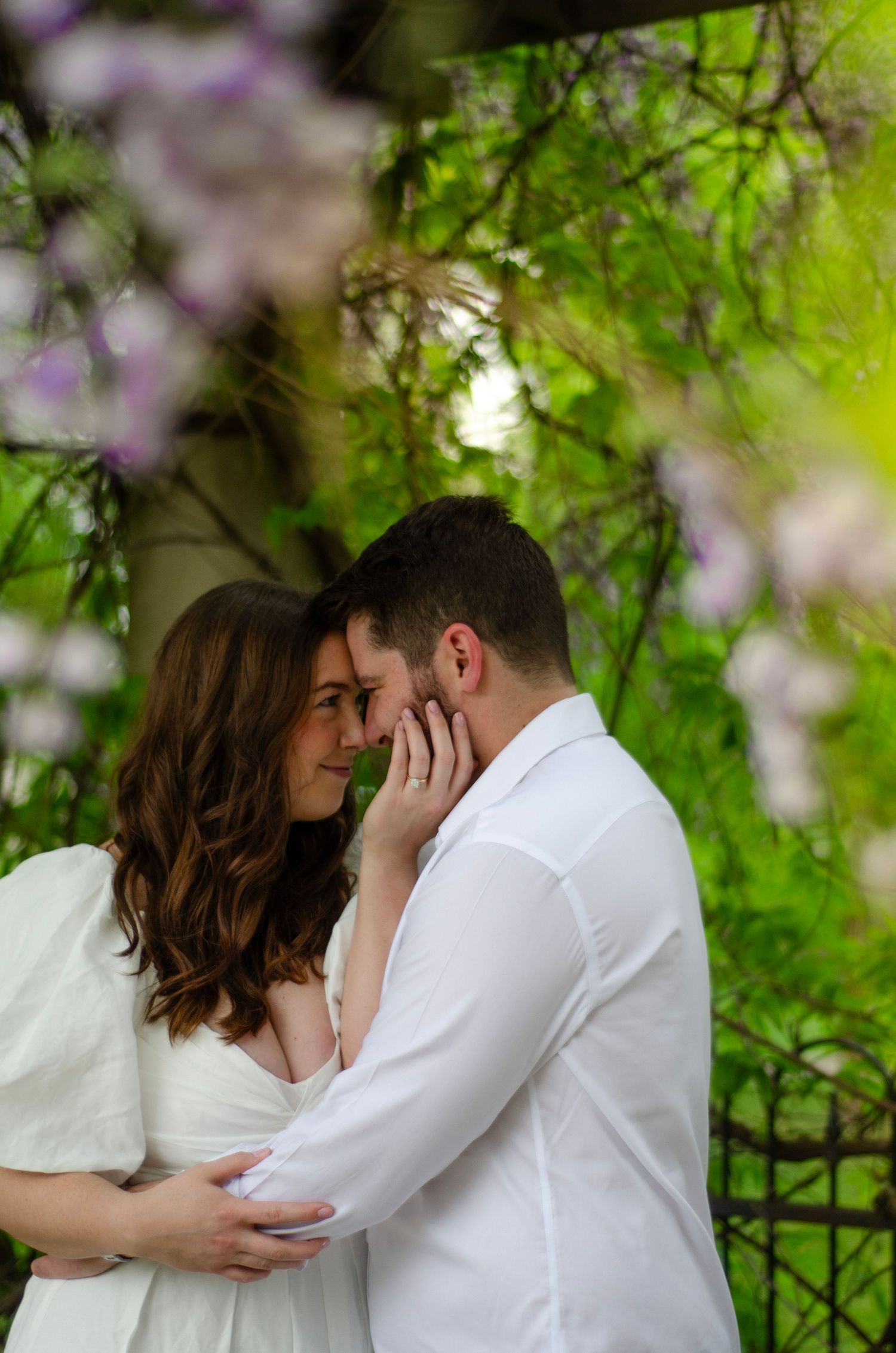 Intimate moment between couple in white attire beneath flowering spring branches.