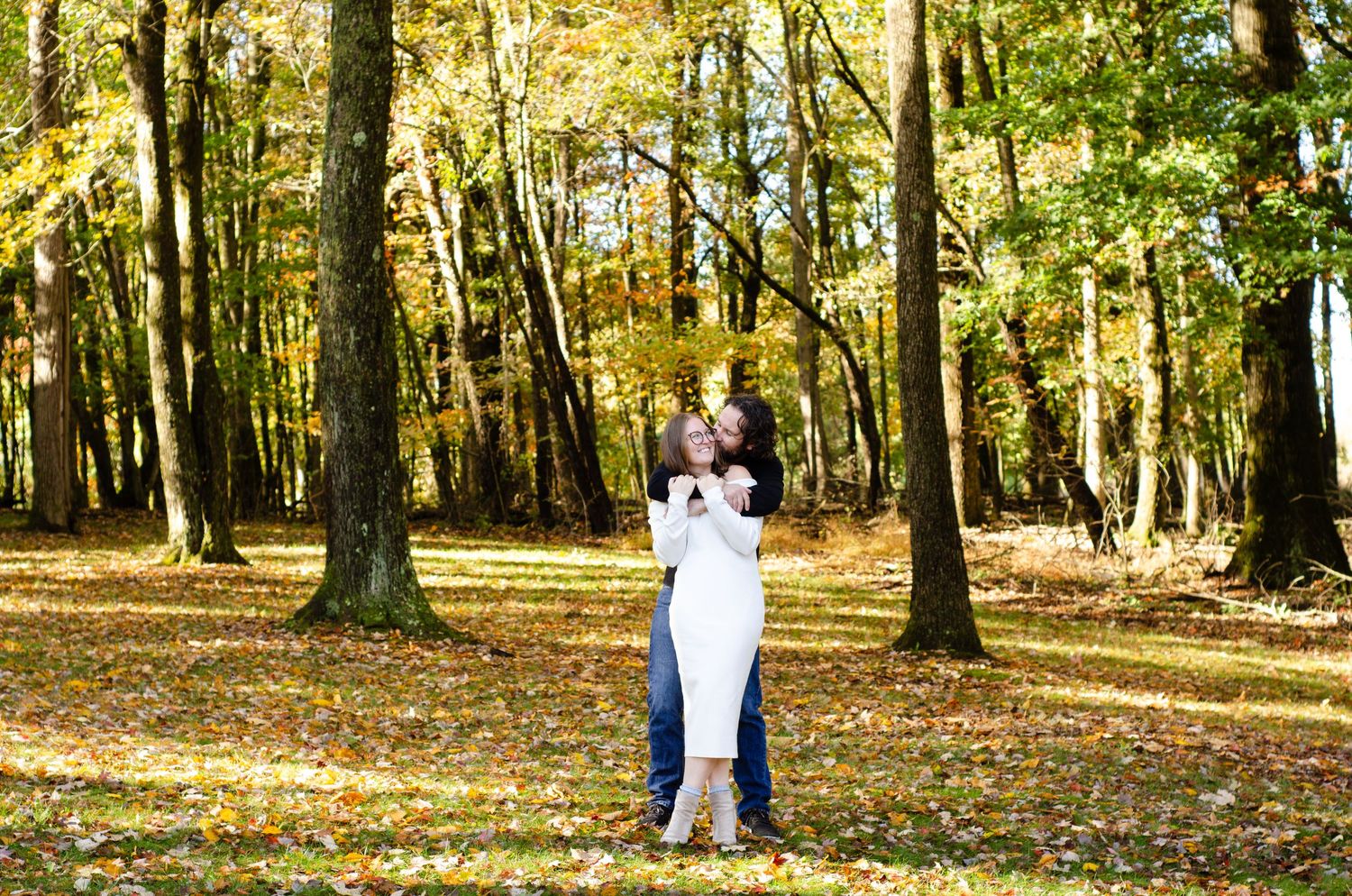 Couple embracing in autumn forest with colorful fall foliage and tall trees in background.
