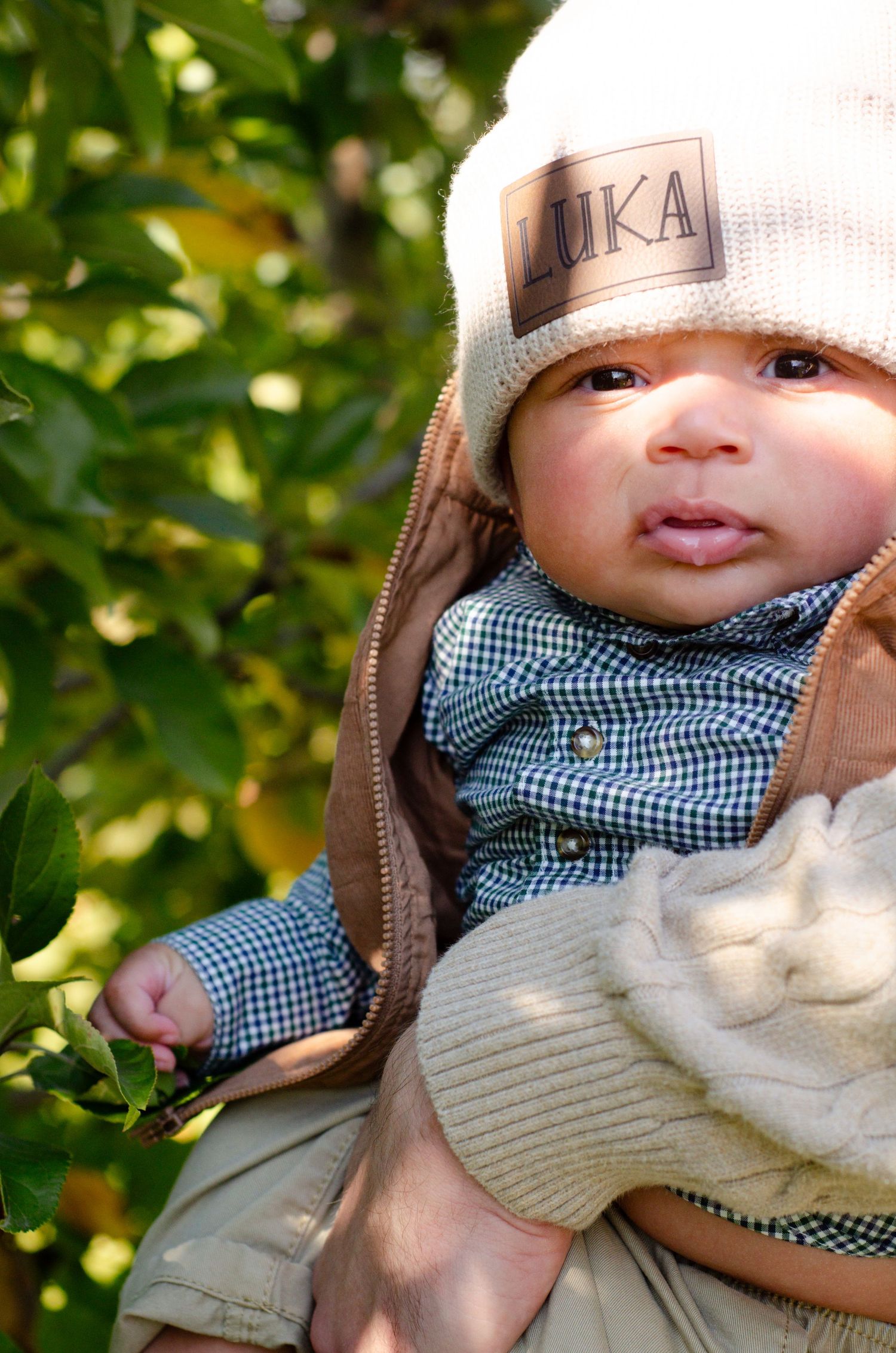 A baby wearing a blue gingham shirt and knit hat smiles at the camera. Family photos by KM Conceptions Photography in Columbus, OH.