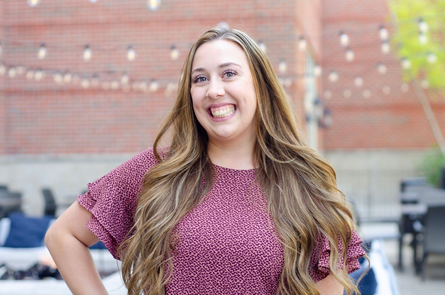 Portrait of person in burgundy top against brick wall with string lights.