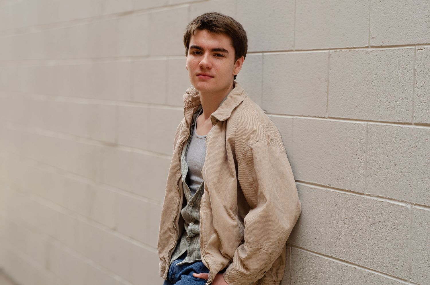A casual portrait against a light gray wall wearing a beige jacket.