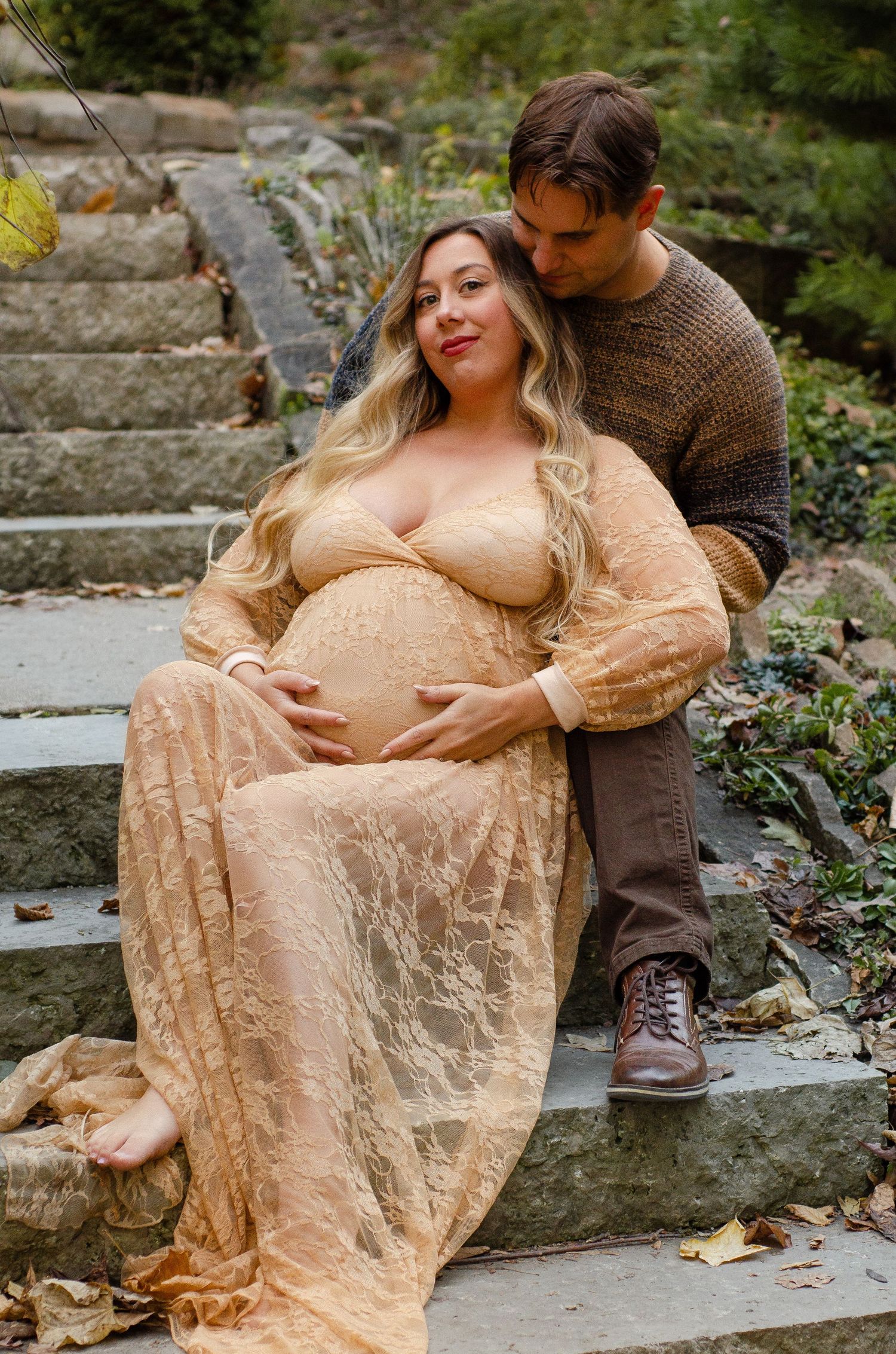 A couple sits on stone steps surrounded by autumn foliage wearing brown and peach tones. Maternity photos by KM Conceptions Photography.
