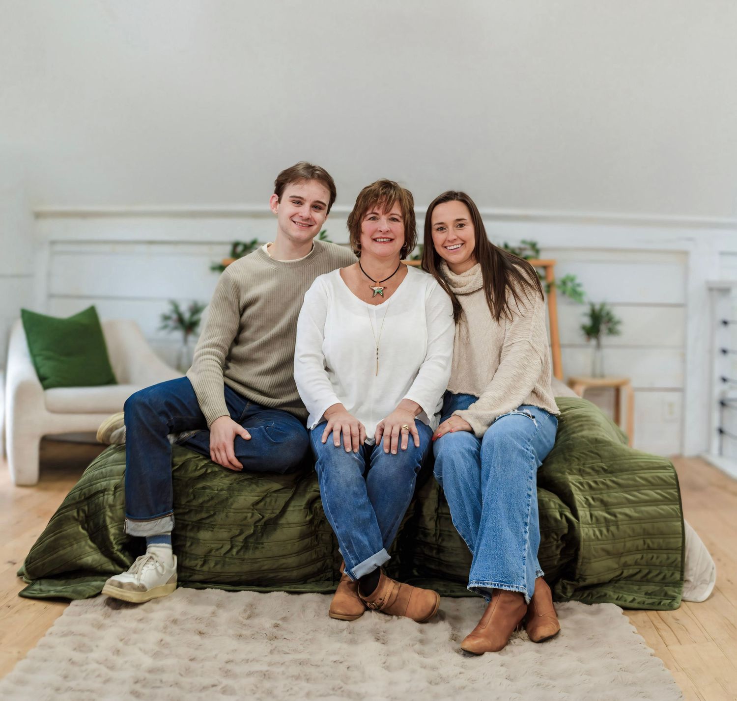 Three people sit casually on a green bedspread in a bright modern bedroom with white walls and decor.