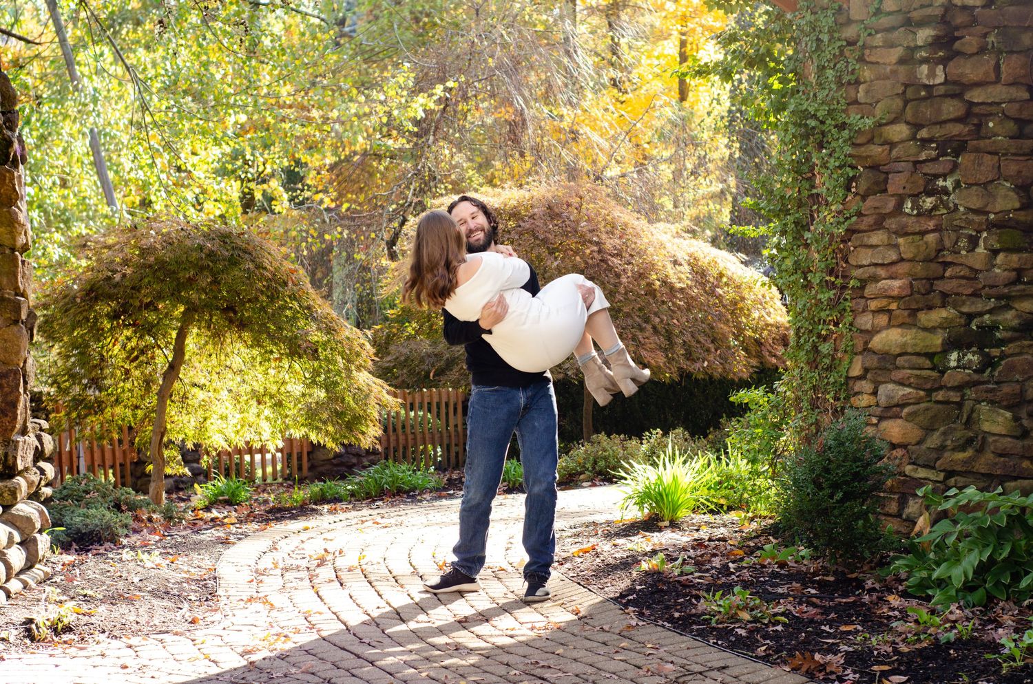 Man lifting woman in garden with autumn trees and stone wall on brick path.