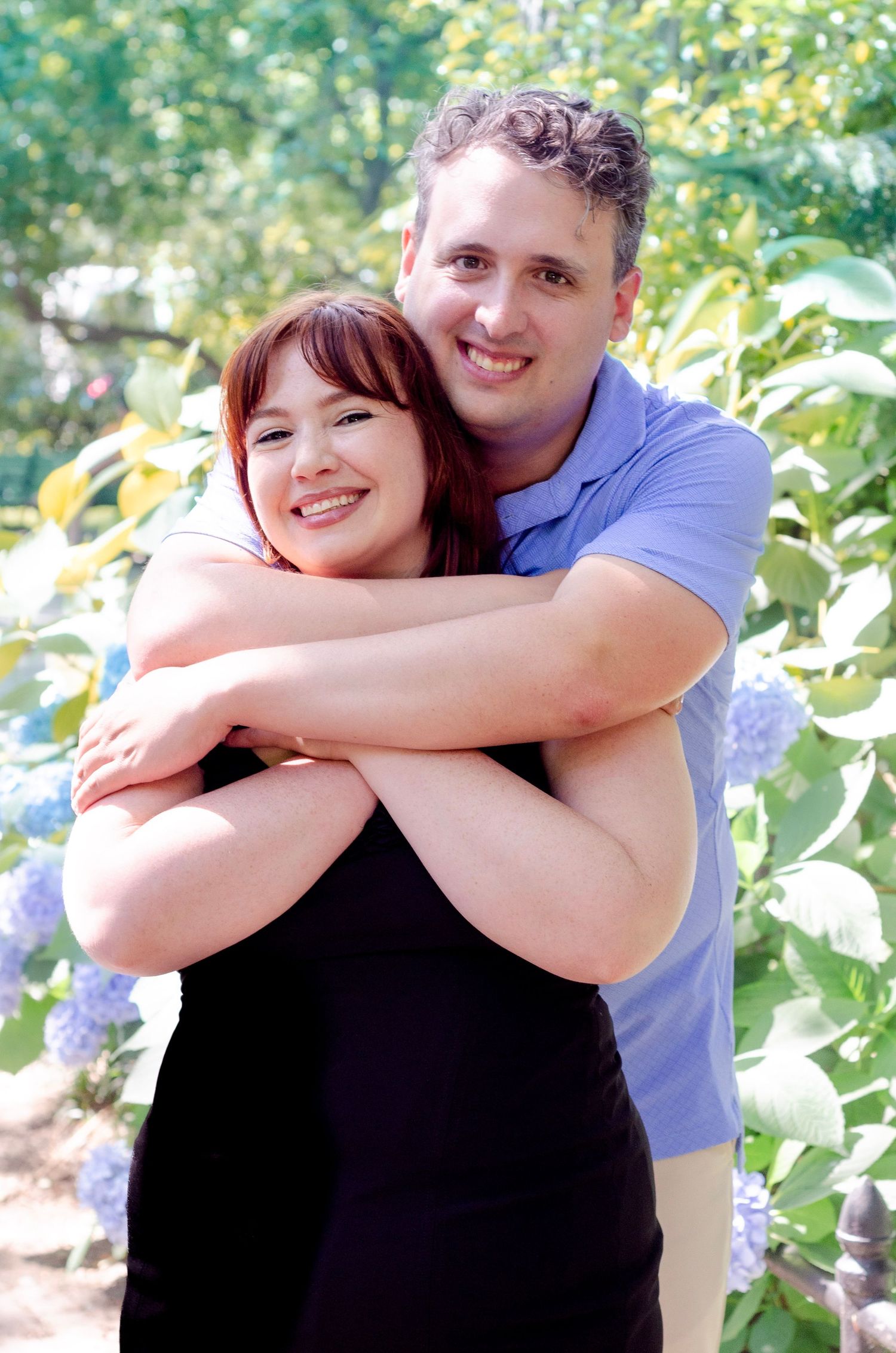 A smiling couple embraces outdoors, with the man hugging the woman from behind near blooming hydrangeas.