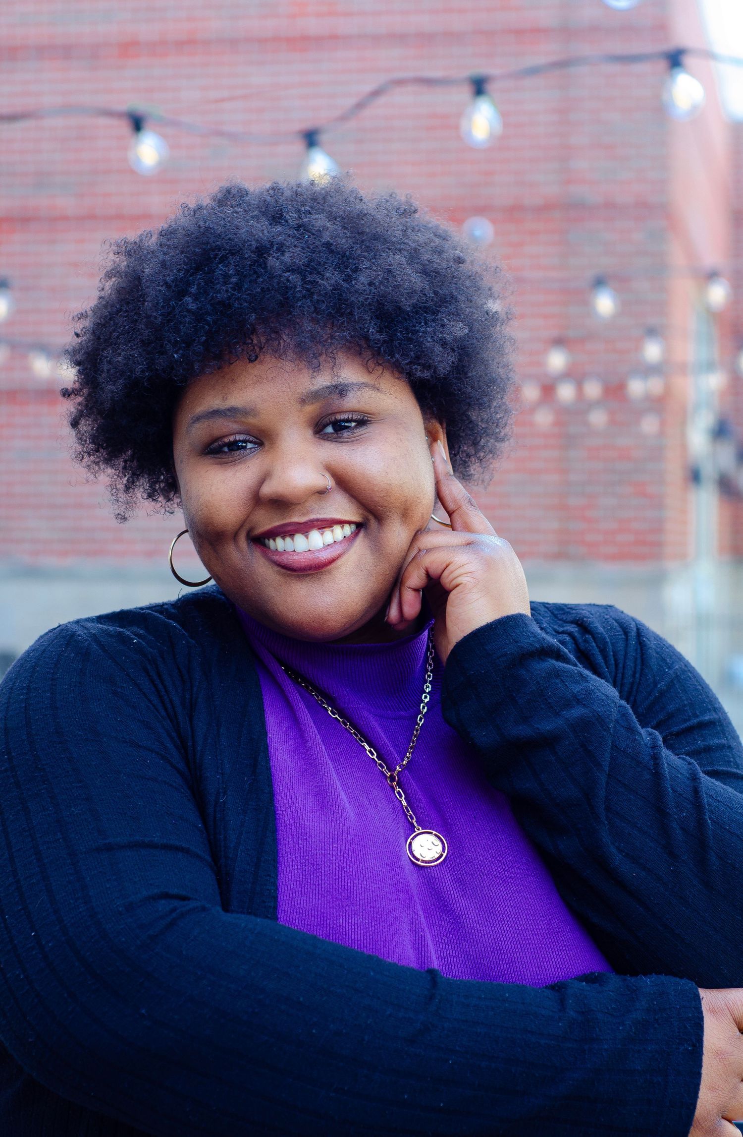 Portrait featuring purple top and black cardigan against brick wall with string lights.
