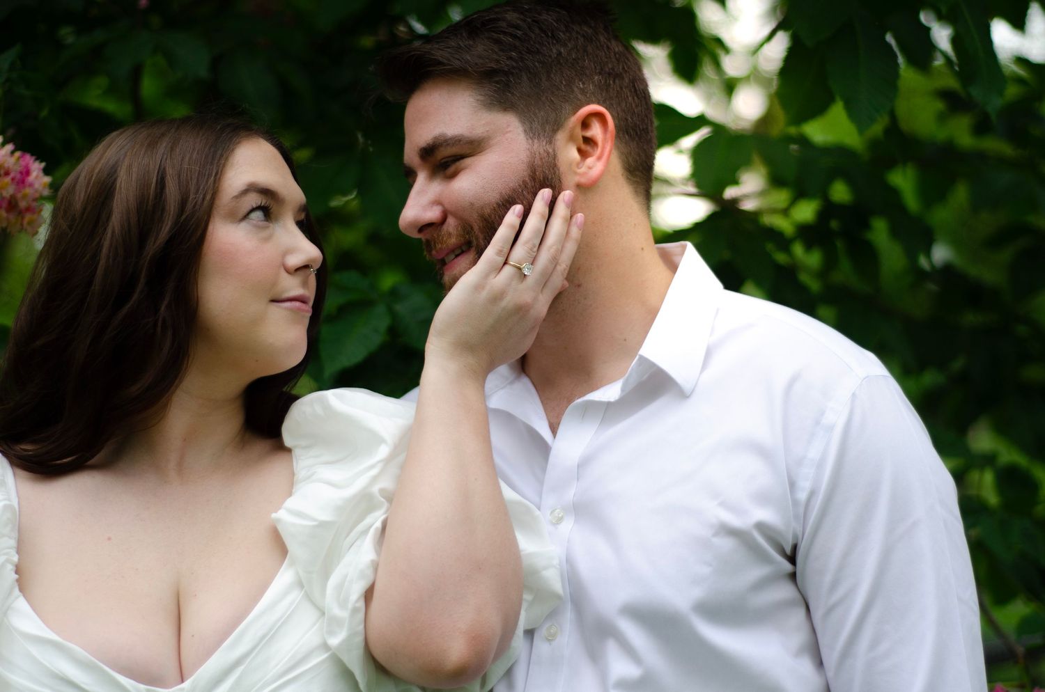 Tender moment between couple in white outfits against green foliage background.