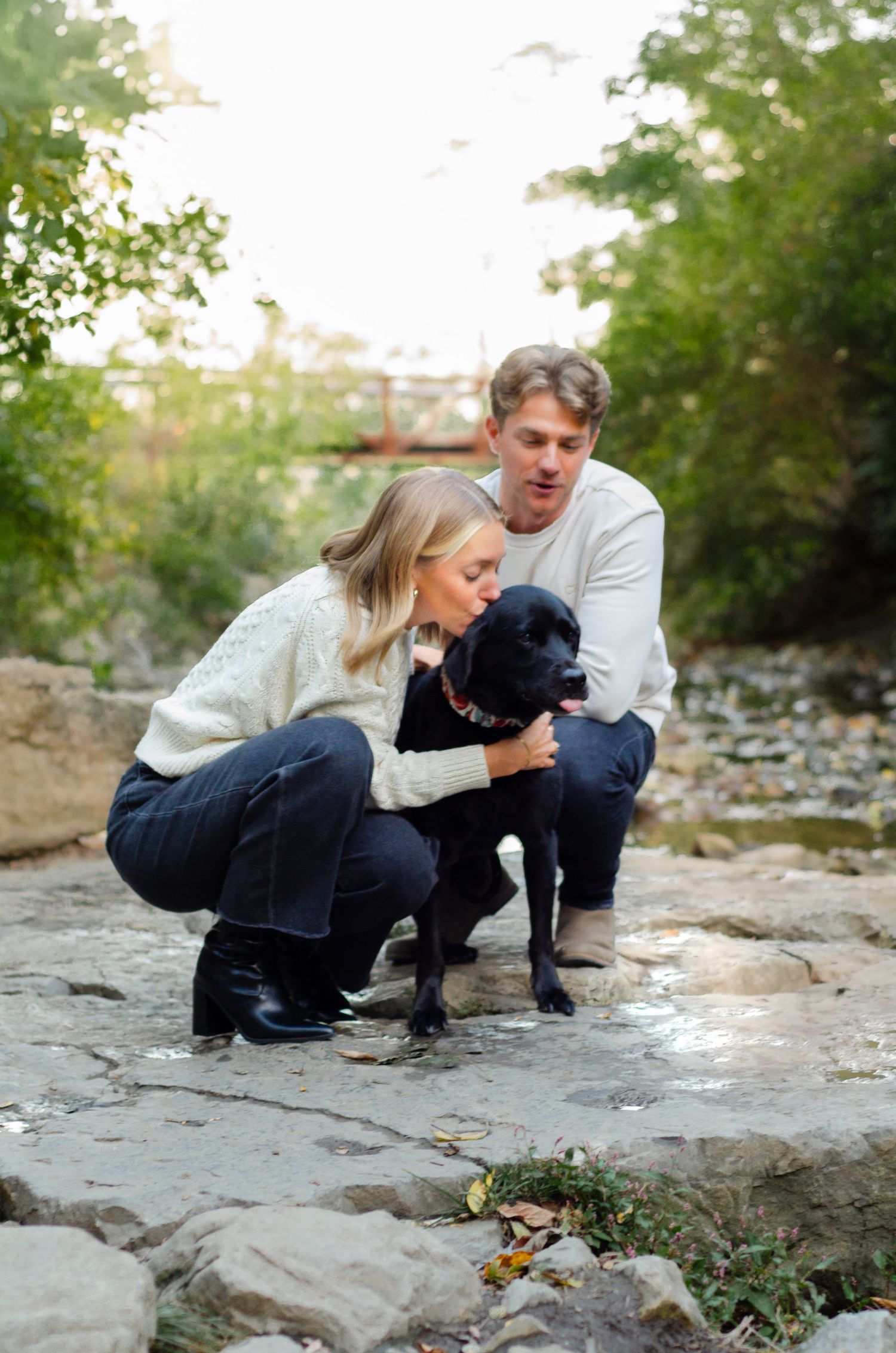Couple crouching outdoors kissing a black Labrador dog on rocky ground surrounded by lush green trees.
