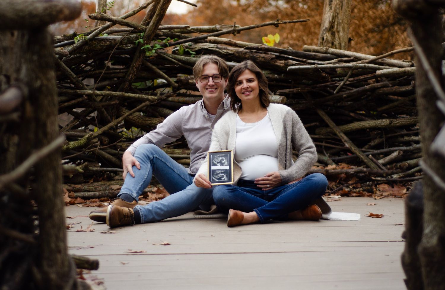A couple sits together on wooden logs while holding ultrasound photos.