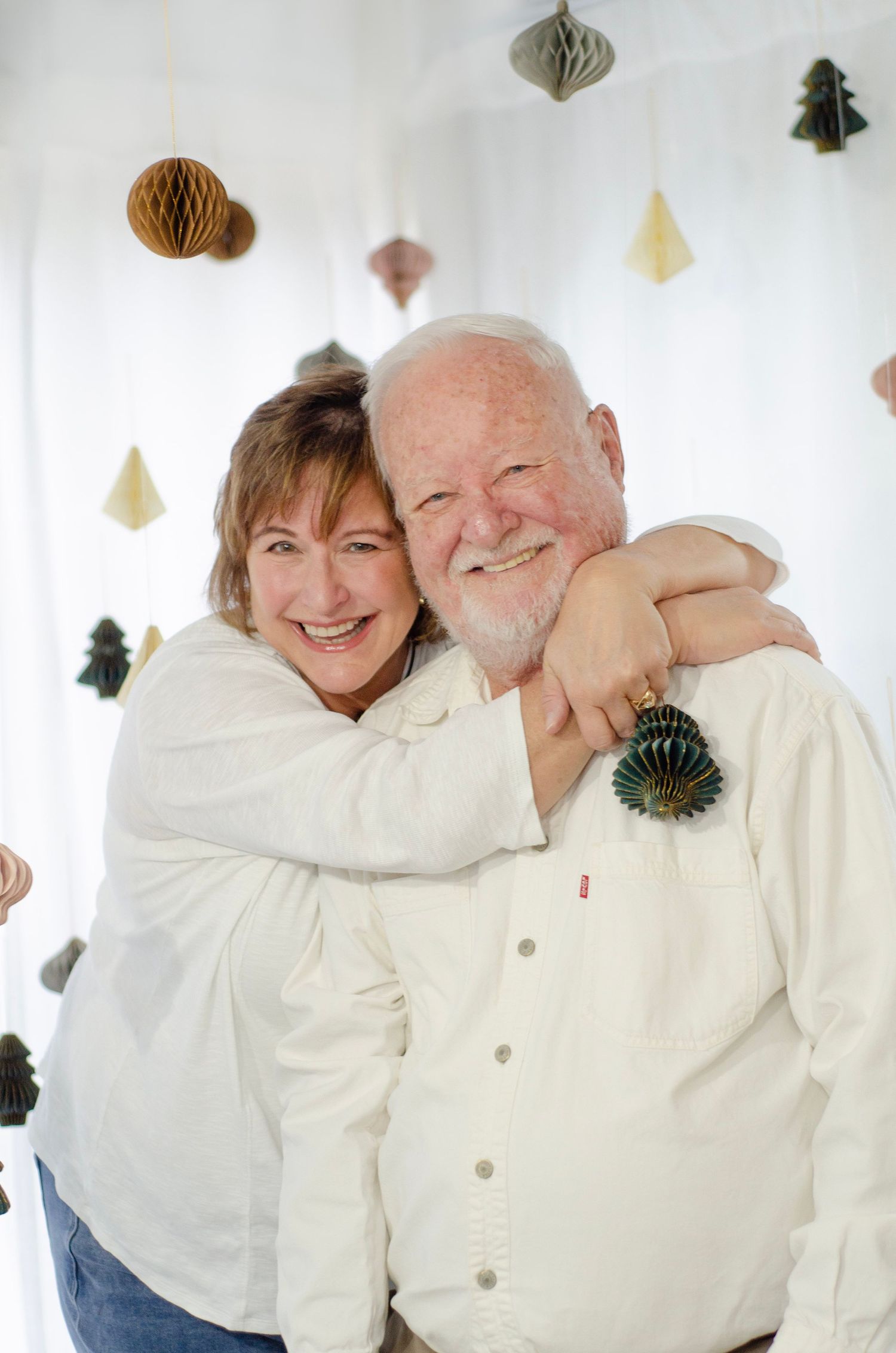 Couple embraces while wearing white clothing against a backdrop with hanging holiday ornaments.