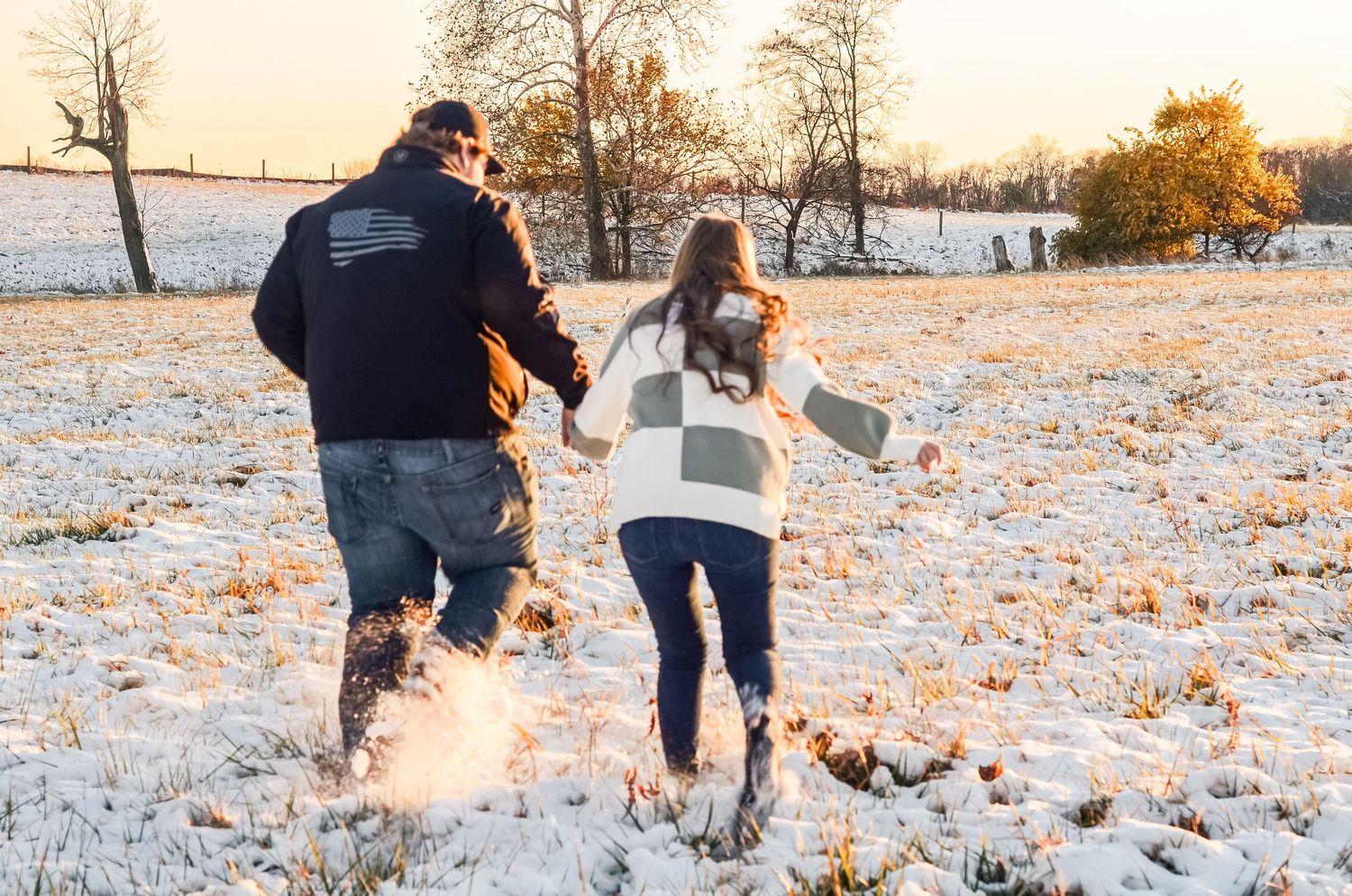 Two people playfully running through a snowy field at sunset with autumn trees in the background.