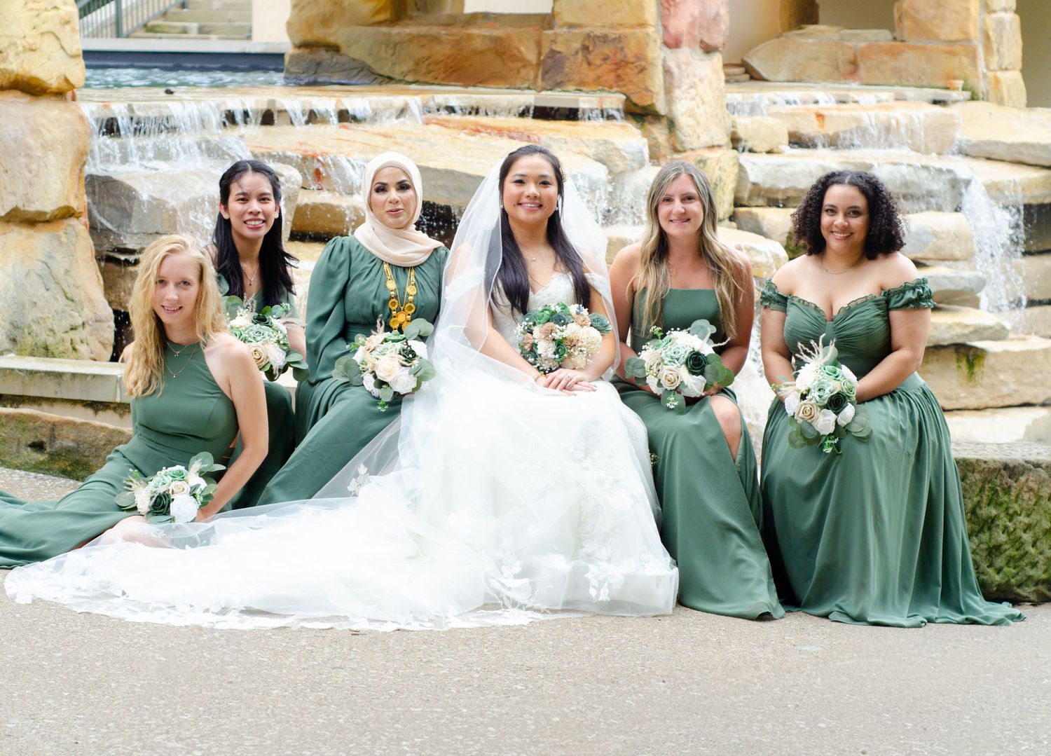 Bridesmaids in sage green dresses sitting on stone steps with white floral bouquets at an outdoor wedding venue.