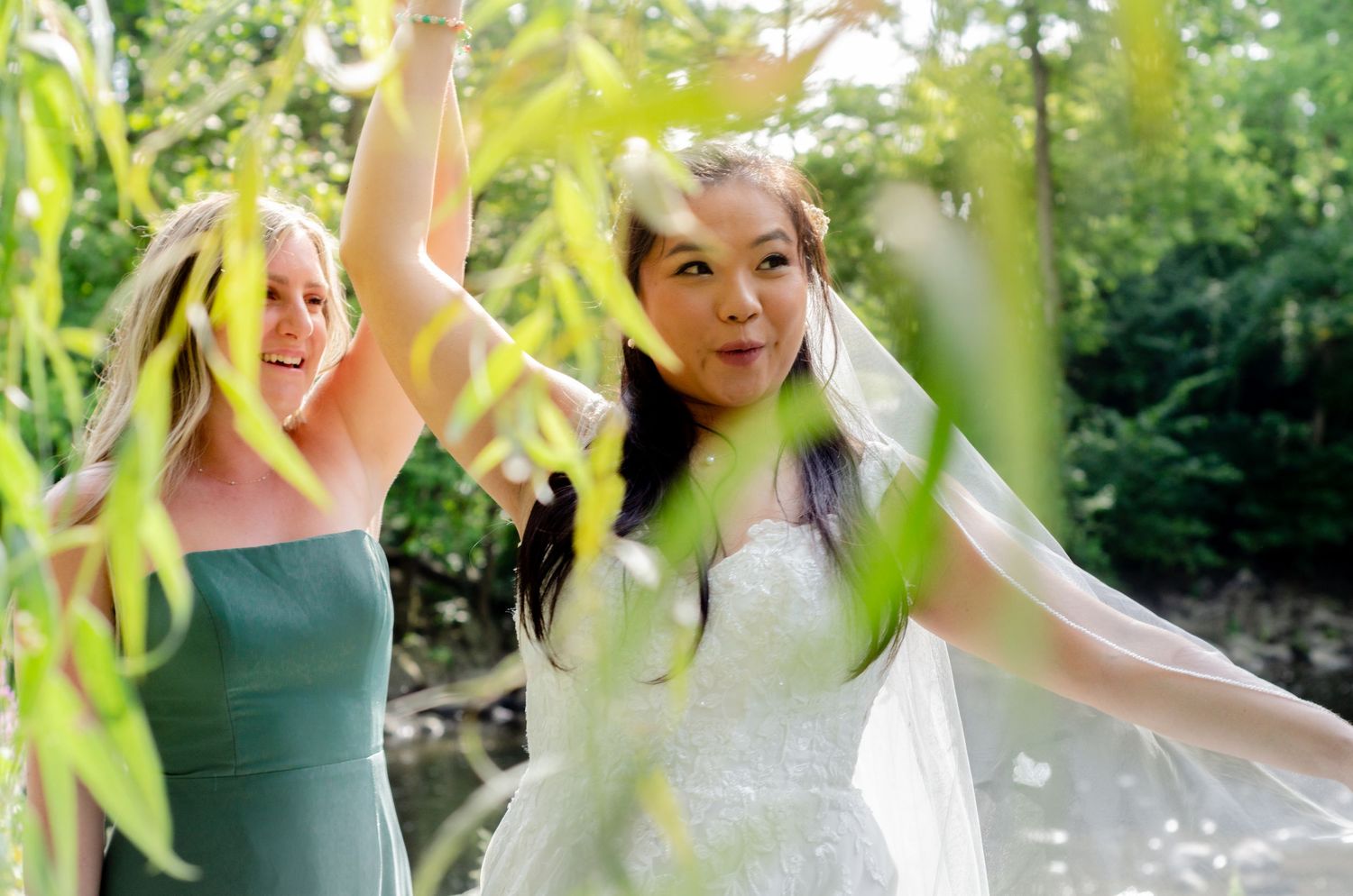 Joyful wedding party members dance and celebrate under hanging willow tree branches in a garden setting.